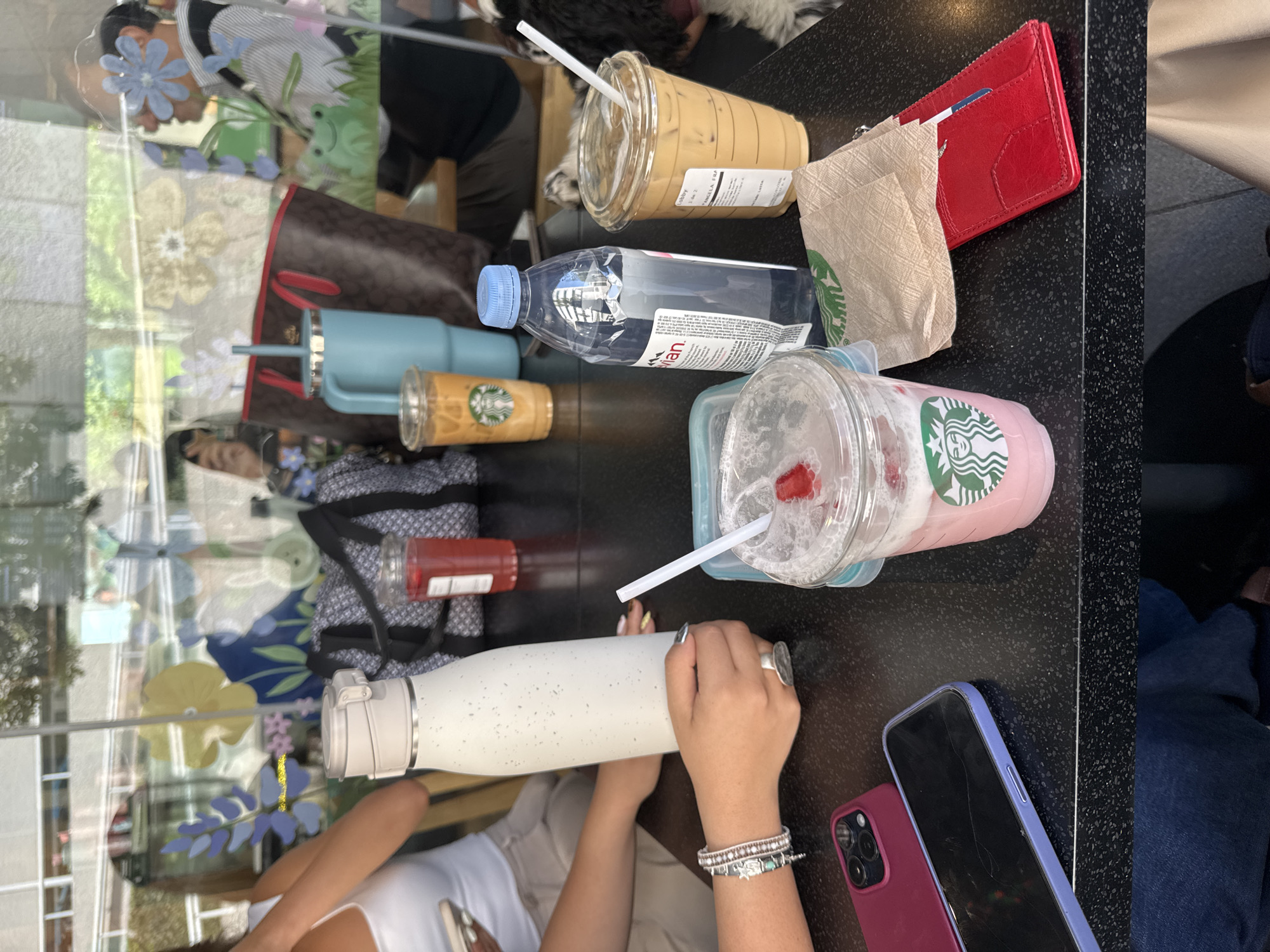 The scene depicts an interior view of a cafe, identified as being in Puebla, Mexico. A dark, speckled tabletop occupies the foreground, laden with various beverages and personal items.
On the table, from left to right:
- Two smartphones are placed horizontally. The leftmost is an iPhone with a maroon case and a triple camera array, face down. To its right, a lavender-cased smartphone lies face up, exhibiting a visibly cracked screen.
- A light-skinned individual's right arm and hand are prominent in the mid-left foreground. The person wears multiple bracelets on their wrist and a silver ring on their finger, holding a white, speckled, insulated water bottle with a light grey cap.
- A transparent plastic Starbucks cup contains a pink, blended beverage with foam, a white straw, and a piece of red fruit floating on top. It sits within a light blue plastic container or lid.
- A clear plastic bottle of Evian water with a blue cap is positioned to the right of the pink drink.
- A crumpled tan Starbucks napkin lies next to the water bottle.
- A red leather wallet or card holder is next to the napkin.
- Another transparent plastic Starbucks cup contains an iced coffee or latte with a white straw and a label on its side.
- A portion of a black and white furry animal, consistent with a dog, is visible on the far right edge of the table, near the iced coffee.
Beyond the table, through a large window acting as a wall, reflections show elements of the cafe's interior and partial glimpses of an exterior environment. Reflected items include:
- A grey and white patterned backpack or duffel bag.
- A large brown tote bag with red handles (Coach brand pattern).
- A light blue insulated tumbler with a straw, next to a clear cup containing an iced orange-colored drink with a Starbucks logo.
- Partially visible individuals include a person in a white tank top, a person with dark hair in profile, and a person in a striped shirt and glasses also in profile, with a backpack strap visible.
The window itself is decorated with large, colorful, stylized floral and nature motifs, including yellow and blue flowers, purple leaves, and a green frog illustration.
The external view beyond the window is blurred, showing distant building structures and hints of greenery, suggesting an urban setting.