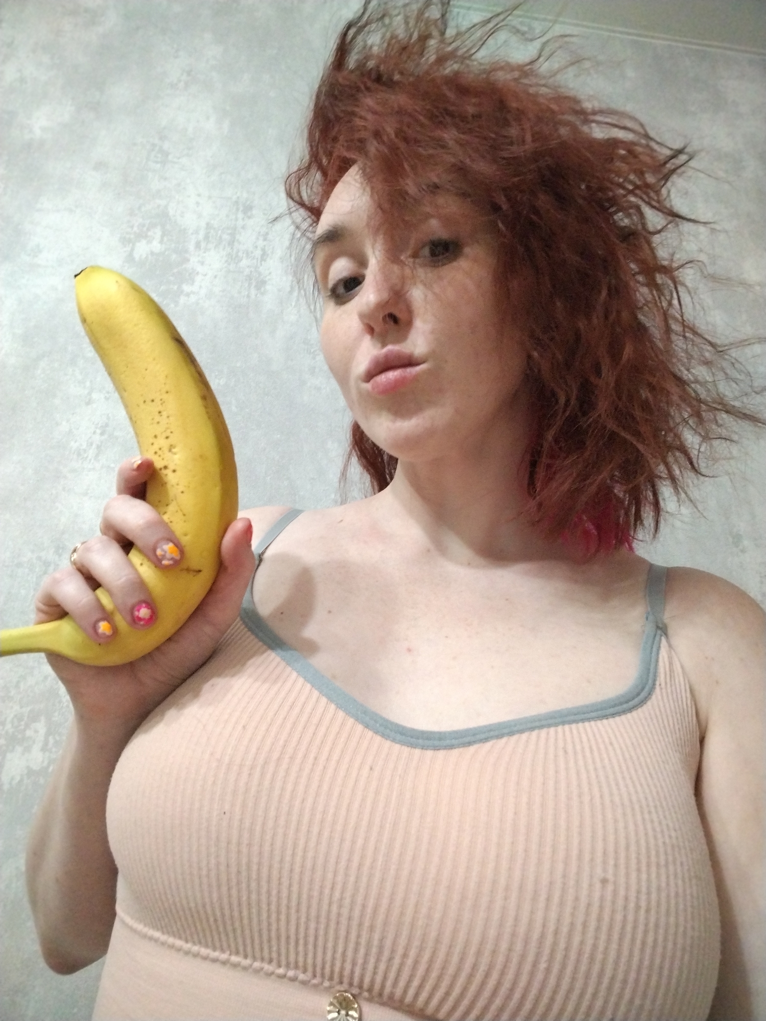 An indoor scene features an adult female holding a banana. The woman has disheveled reddish-brown hair with pink highlights and light skin with visible freckles. She looks directly at the camera with a pursed-lip expression. She wears a light peach-colored ribbed top with grey trim and straps; a decorative gold-colored button is on the lower front. Her left hand, displaying a ring on the ring finger and multi-colored floral nail art (orange, pink, and white), grips a ripe, yellow banana with minor dark spots, holding it vertically in front of her torso. The background consists of a plain, light grey textured wall. The precise location within Izhevsk, Russia, is not visually ascertainable from the image; it appears to be a residential interior.