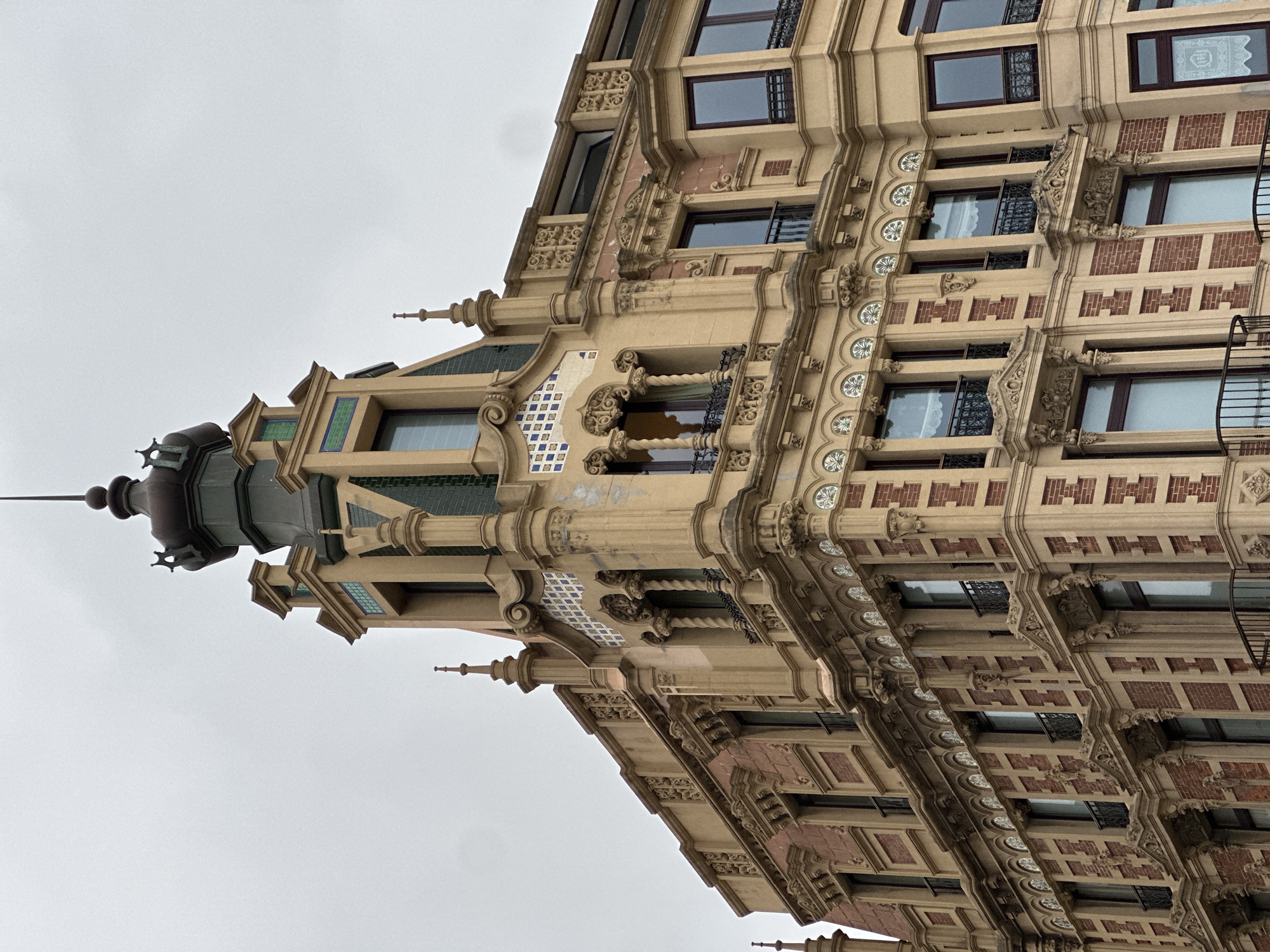 The image shows an upward-angled view of a building's facade, likely located in Gijón/Xixón, Spain, due to the distinctive architectural style. The building features ornate detailing, including decorative moldings, patterned brickwork, and a series of evenly spaced windows. A turret-like structure tops the building, adorned with a pointed spire and further embellishments. The sky is overcast, contributing to a muted color palette. There are no people visible in the frame. The scene focuses on the architectural details of the building.