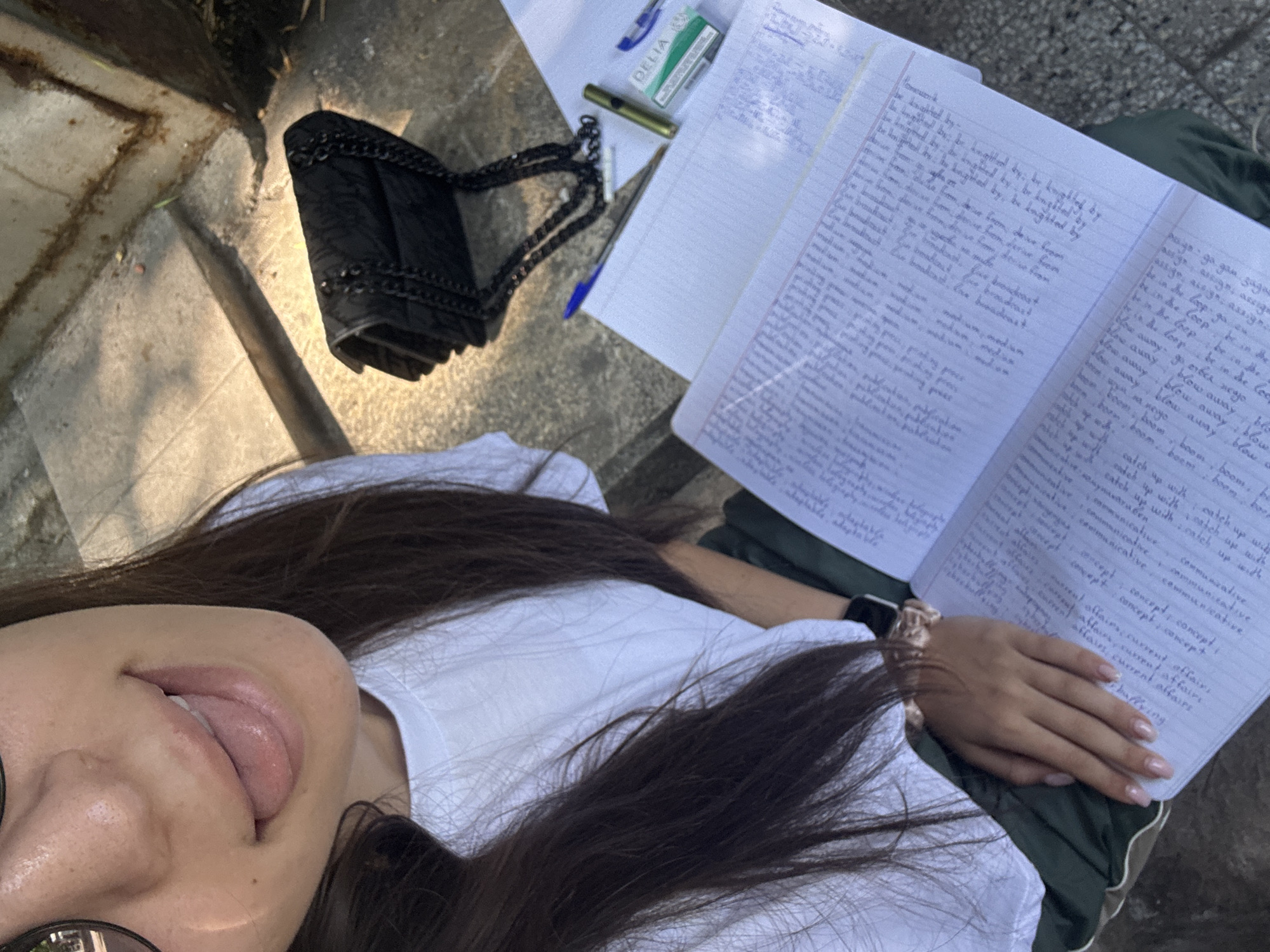 A high-angle, close-up shot captures a person outdoors in Plovdiv, Bulgaria, under bright daylight. The subject, visible from the chin to the torso, has long dark hair, wears a white t-shirt, and sunglasses. Her tongue is slightly visible, suggesting a candid moment. On her right wrist is a scrunchie.
The person is positioned next to an open, lined notebook filled with handwritten English vocabulary and phrases. On the left page, text includes "Homework," followed by lists containing "be knighted by," "drive from," "live broadcast," "medium," "printing press," and "publication." The right page lists terms such as "assign," "be in the loop," "blow away," "boom," "catch up with," "communicative," "concept," "current affairs," and "buffering."
Adjacent to the notebook are a black quilted purse with a chain strap, a small green box labeled "DELIA," a gold-colored item (possibly a pen or small lighter), and a blue ballpoint pen. The setting appears to be a casual outdoor area, possibly concrete steps or a raised platform, with some indistinct greenery or ground visible in the background.
