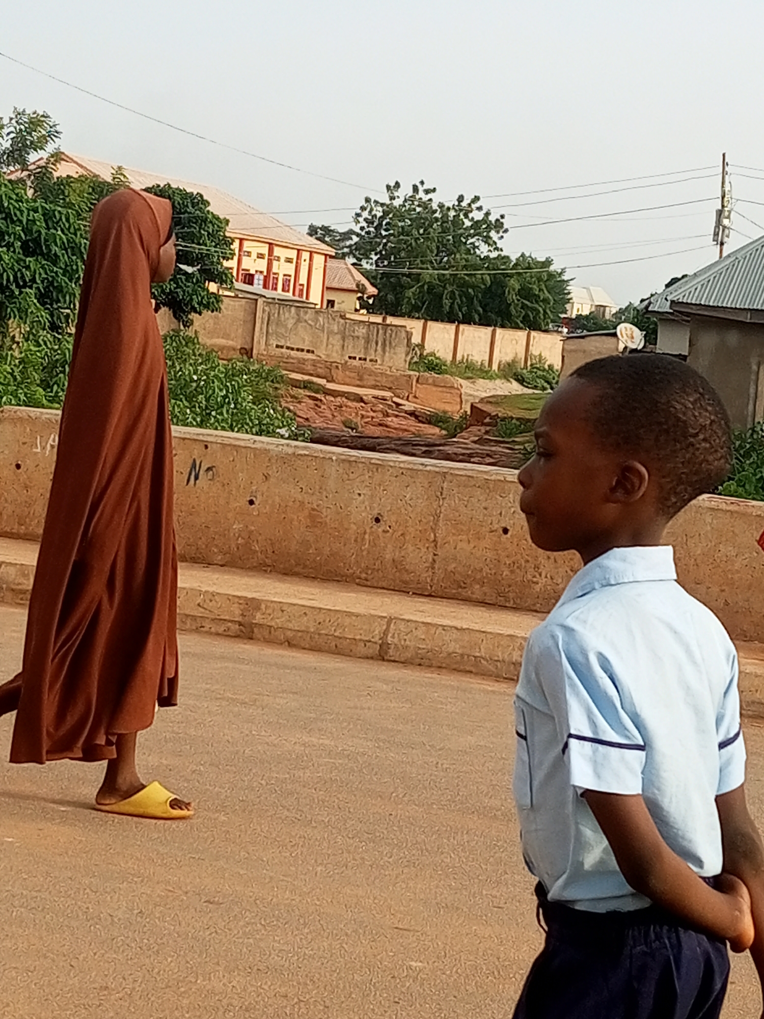 In the city of Tumfure, Nigeria, a young boy in a light blue school uniform, with his hands clasped behind his back, walks on a paved road. He is looking to his left. To his left, a person wearing a brown hijab and yellow slippers is walking away from the camera. The road is bordered by a concrete barrier, and beyond that, there are trees, buildings, and power lines visible. The sky is overcast.