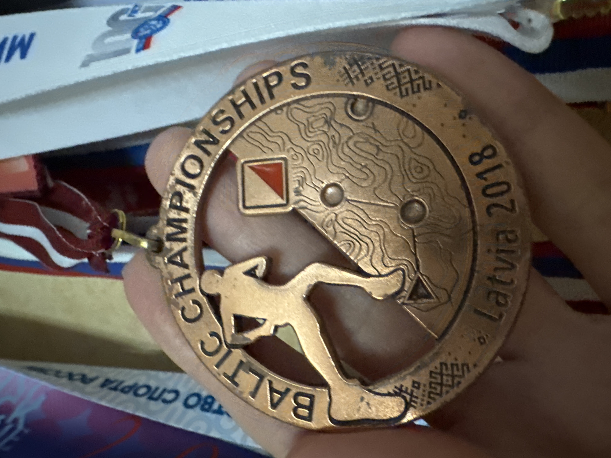 The image shows a hand holding a bronze medal. The medal has a runner silhouette on it and reads "BALTIC CHAMPIONSHIPS LATVIA 2018". The background shows parts of other medals and ribbons. The location of Sinyavino, Russia is not depicted or implied in this image.