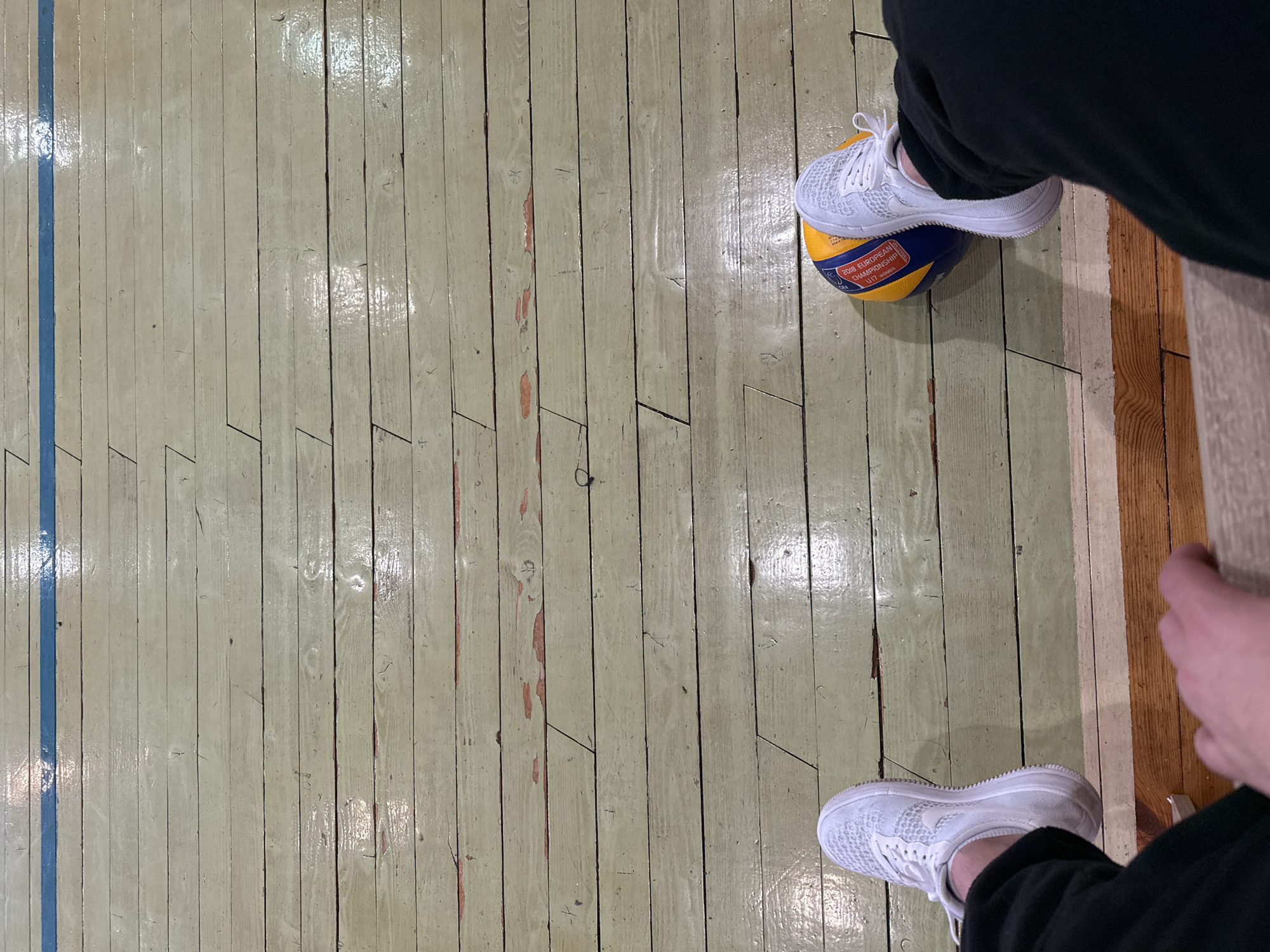 The scene is a top-down view of a person standing on a wooden floor in a gymnasium. The gymnasium is likely located somewhere in Sofia, Bulgaria but the exact location is unknown.
The floor has light green-grey painted wooden planks with a visible blue line running along one side. Some of the planks show signs of wear and tear, with chipped paint revealing the wood underneath.
The person is wearing black pants and white athletic shoes. One foot is resting on a volleyball, compressing it slightly. The edge of a wooden bench or other structure is visible to the right, with a hand resting on it.