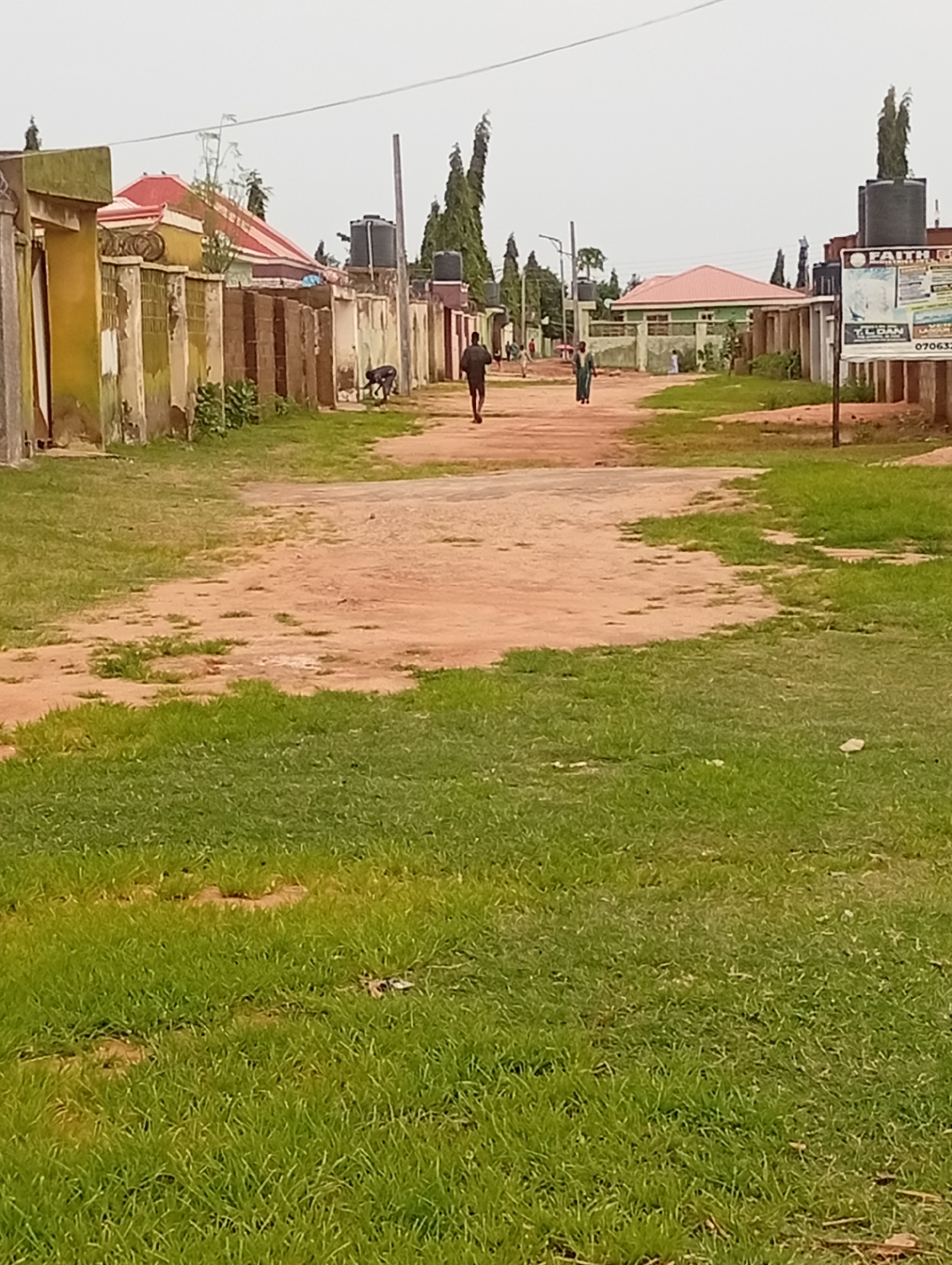 The image shows a dirt road in Tumfure, Nigeria, lined with buildings and vegetation. In the foreground, a grassy area with patches of bare earth is visible. The road itself is dusty and appears to be a pathway through a residential or commercial area. Several people are seen walking on the road. On the left side of the road, there are buildings with fences. One building has a yellow facade and a red roof. Further down, there are other structures with varying architectural styles and water tanks on some of them. On the right side of the road, more buildings are present, some with fences, and a large billboard is mounted on a pole. Tall, slender trees are scattered throughout the scene, especially towards the background. The sky is overcast.