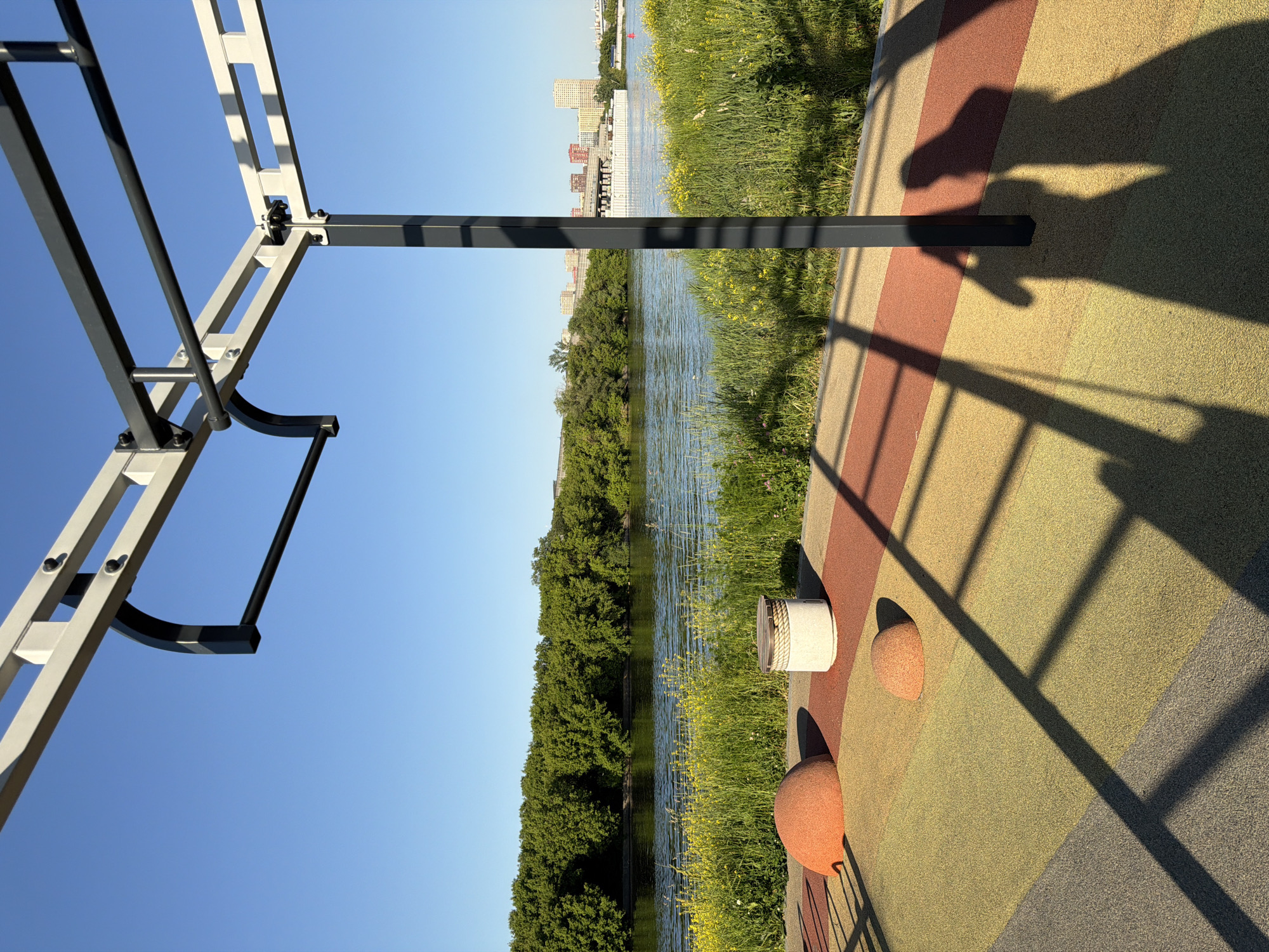 This image depicts an outdoor fitness area situated on the embankment of the Moskva River in Moscow, Russia, specifically within a recreational park on Mnyovniki Island. The perspective looks southeast across the river towards the Fili-Davydkovo district.
In the foreground, a modular metal fitness structure, primarily light grey with dark grey/black horizontal bars and supports, occupies the upper part of the frame. The ground beneath is covered with a multi-colored EPDM rubber safety surface, segmented into dark grey, light yellow-green, and reddish-brown/terracotta sections. Embedded within this surface are two small, hemispherical, reddish-orange rubber mounds and one cylindrical, off-white structure topped with decorative rope, likely functioning as a seat or obstacle. Long, sharp shadows of the fitness structure and two human figures (one likely the photographer) are cast across the colorful ground towards the lower right, indicating low-angle sunlight from the left, suggesting either early morning or late afternoon.
Immediately beyond the fitness area, a narrow strip of vibrant green grass densely populated with yellow wildflowers (such as dandelions or buttercups) borders the Moskva River. The river's surface is calm with minor ripples, reflecting the clear blue sky.
On the opposite bank, a continuous, dense treeline of mature green deciduous trees forms a substantial natural barrier. In the distant background, to the right of the treeline, an urban skyline emerges, featuring several modern high-rise residential buildings, some with distinctive reddish-orange facades (consistent with the Filigrad complex). A bridge structure, likely a railway or road bridge, spans the river in the middle distance. A large, flat, white vessel, possibly a construction barge or pontoon, is visible on the river in front of the distant buildings. A small red buoy marks a point on the river's surface to the right of the white vessel. The sky above is uniformly clear and bright blue.