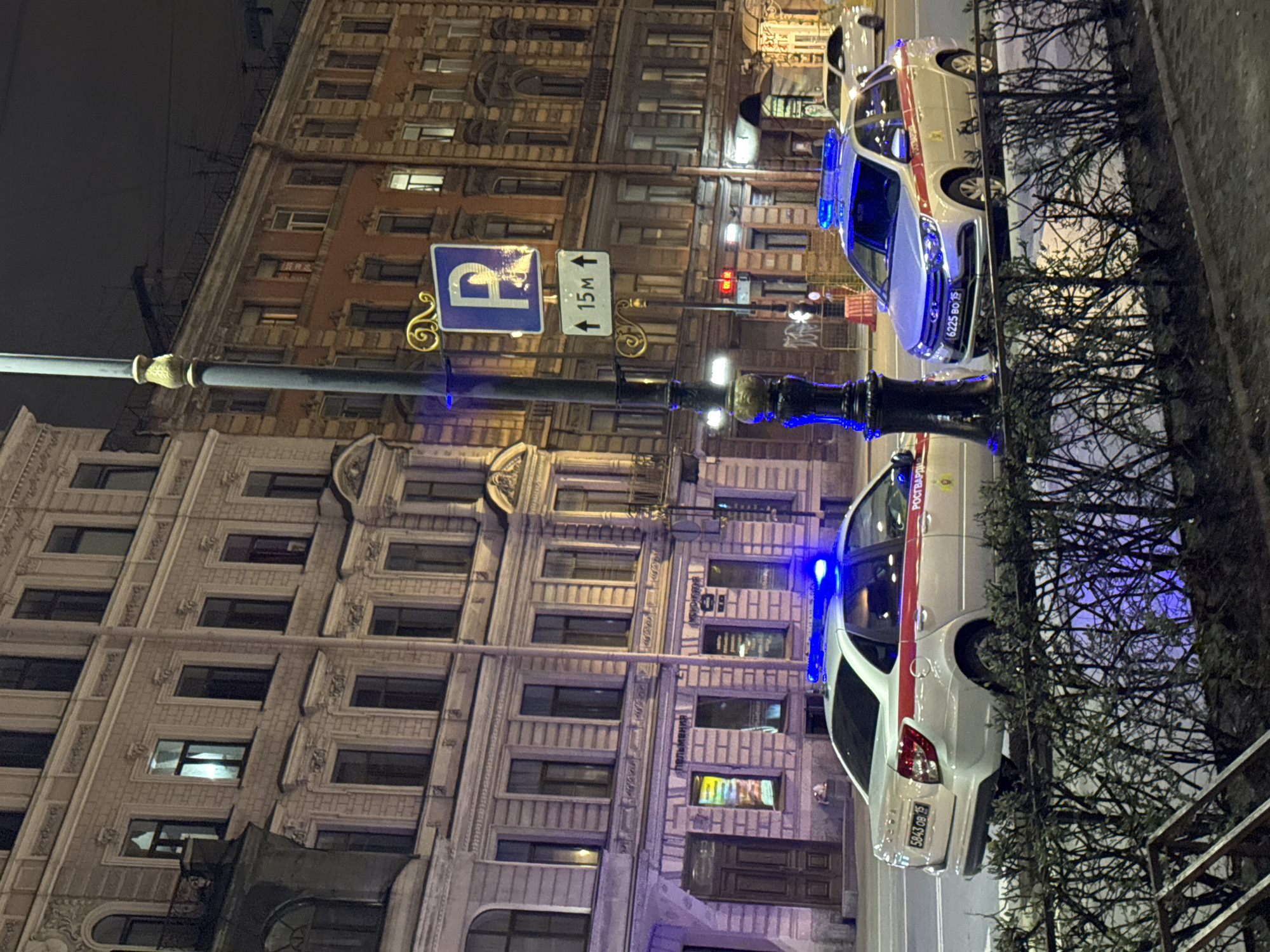 The image depicts a street scene in Saint Petersburg, Russia, at night. Two police cars are parked along the curb. The vehicles are white with a red stripe and have blue emergency lights activated. The license plate of the closer car is visible, reading "5043 BO 98."
In the background are several multi-story buildings, illuminated by streetlights and light emanating from within the structures. A street sign, indicating parking with a directional arrow and a distance of "15M," is attached to a decorative streetlamp. The environment appears to be urban, with a sidewalk or curb along the edge of the street. There is a dense thicket of bushes along the edge of the sidewalk.