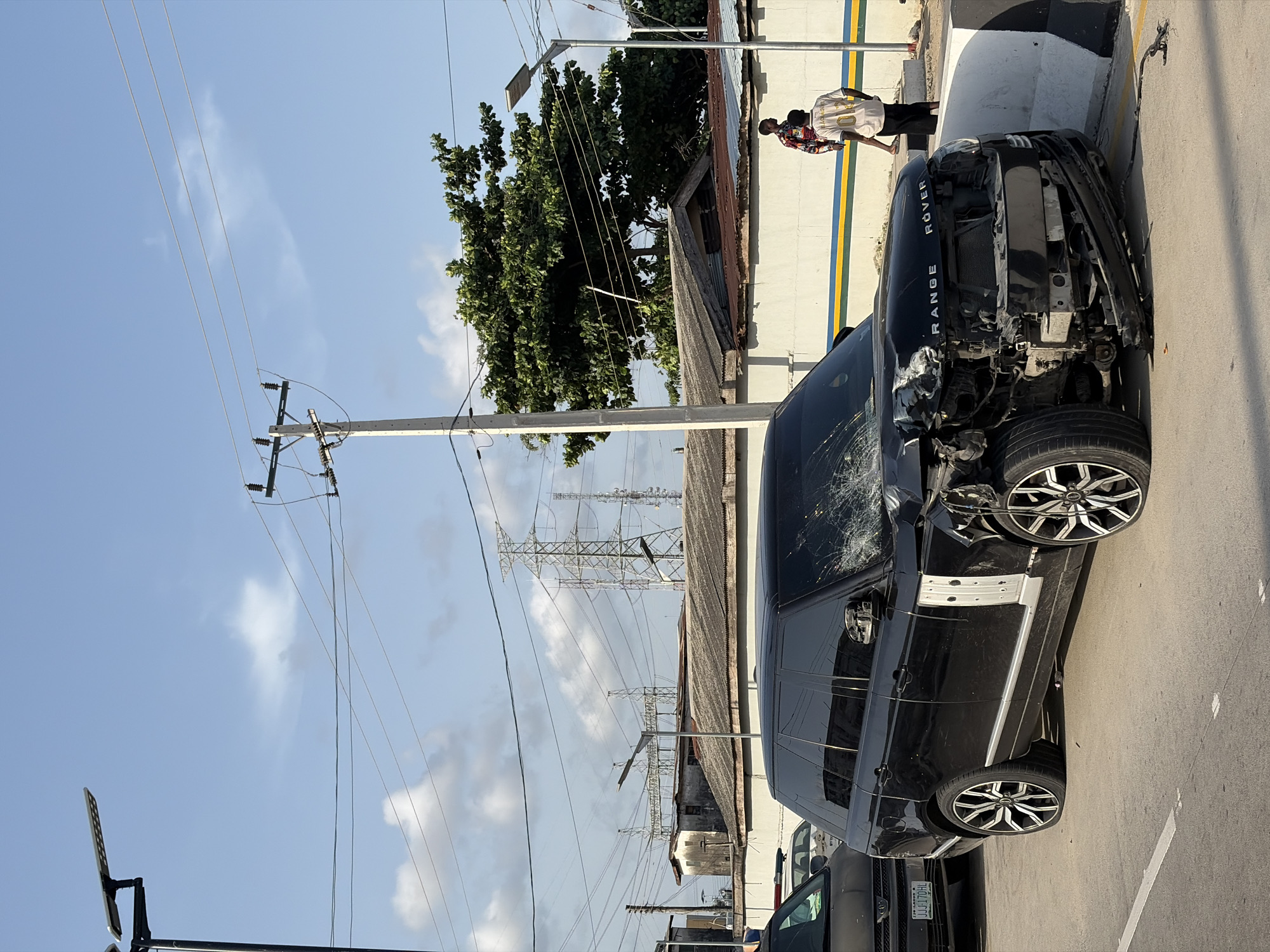 The image shows a damaged black Range Rover on a street in Igboefon, Nigeria. The front of the car is severely damaged, with missing parts and exposed internal components. The windshield is cracked. The car is parked near a sidewalk and a building with a white wall and yellow and blue stripes. Two people are standing near the front of the damaged vehicle. Overhead, there are power lines and poles. Other vehicles are parked nearby, and high-tension power line towers are visible in the background. The sky is partly cloudy.
