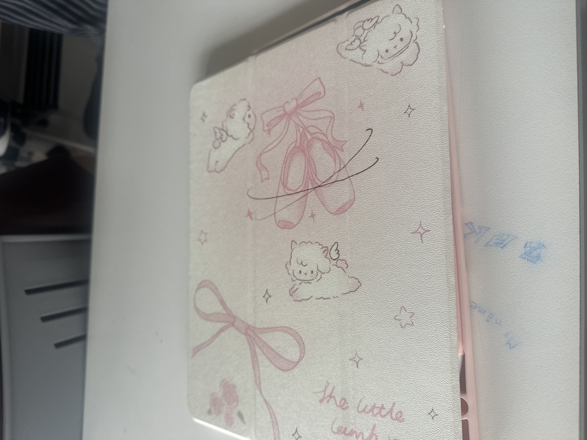 The image shows a close-up of a tablet case. The case is white with pink designs, including ballet slippers with a bow, cartoon lambs with wings, bows, stars, and text that reads "the little lamb". The case is resting on a white surface. To the left of the case, there are glimpses of what appears to be a white storage unit with horizontal slits. The background reveals part of a striped garment. The inscription “My Name” in blue ink can be seen on the white surface to the right of the cover. The image does not contain any discernible information about the location in the city of Chongqing High-tech Zone, China.