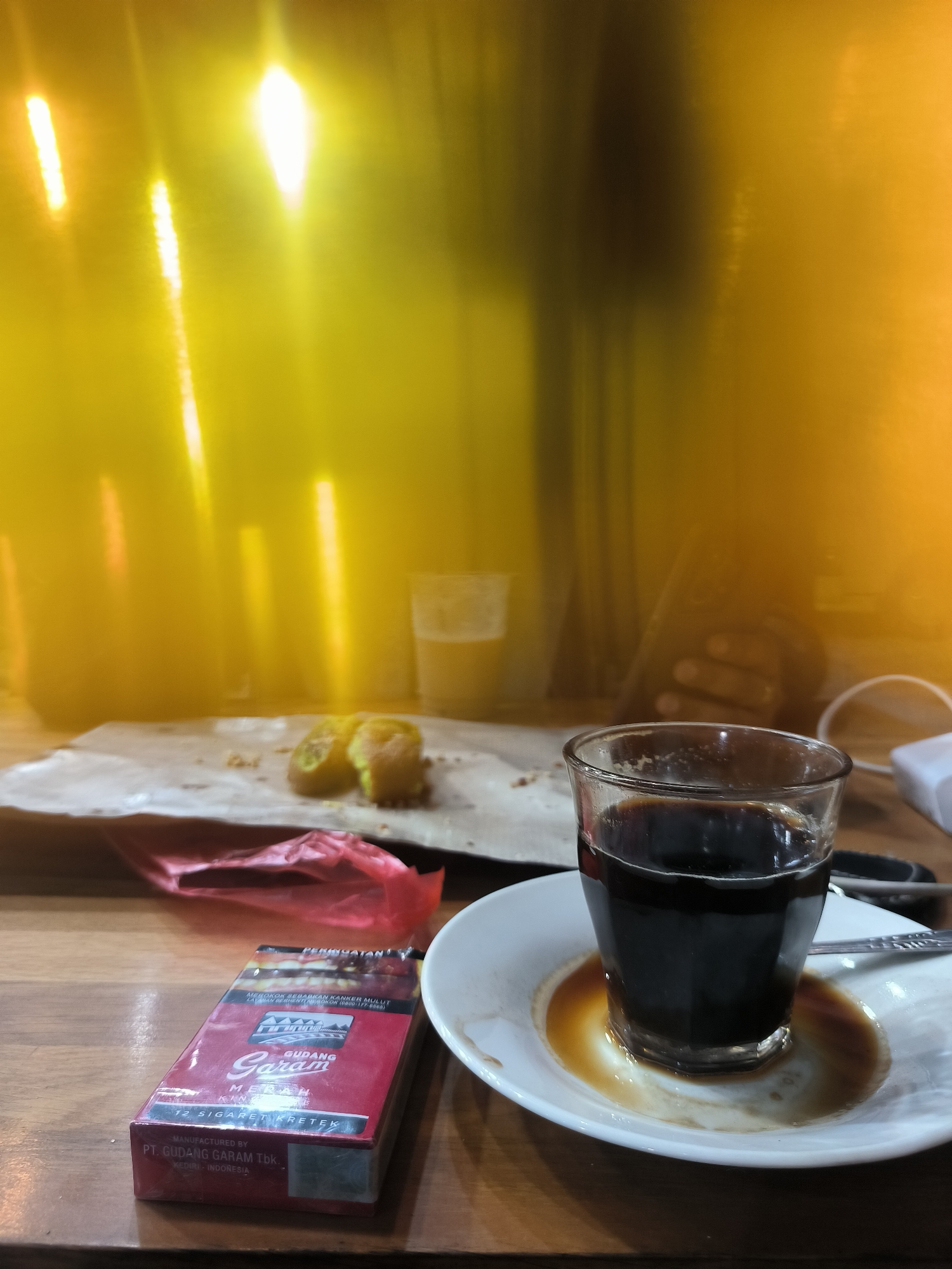 A dimly lit indoor scene features a wooden table on which are placed a glass of dark coffee on a saucer, a red pack of Gudang Garam kretek cigarettes, a crumpled red plastic wrapper, a piece of food on a sheet of paper, and another glass of liquid. In the background is a blurry figure of a person holding a device, possibly a smartphone, in their hand, and a white electronic device. The lighting is diffused, with streaks of yellow and orange.