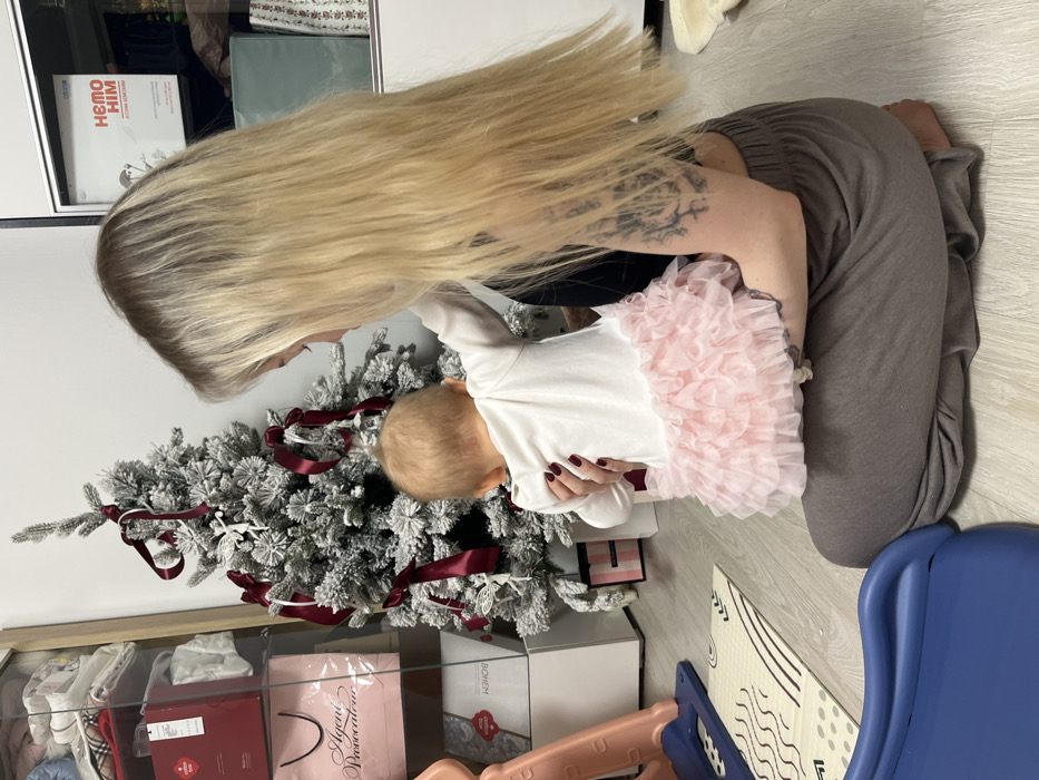 A woman with long blonde hair kneels next to a Christmas tree, assisting a baby in a white onesie with pink ruffles. The tree is decorated with silver ornaments and burgundy ribbons. The woman wears grey pants and a dark top. Underneath the tree, there are gift bags. A blue and beige patterned play mat and a blue plastic object are on the floor beside them.