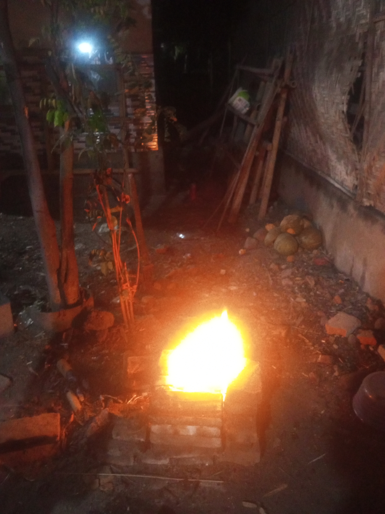An outdoor, nighttime scene in Panggungan, Indonesia, depicts a fire burning in a makeshift brick furnace. The fire is bright orange and yellow, illuminating the surrounding area. To the left of the furnace are several tree trunks, one of which is partially obscured by foliage. In the background, a wooden ladder leans against a textured wall, and a pile of round, brown objects, possibly gourds or coconuts, sits on the ground. The ground itself is a mixture of dirt, small stones, and scattered debris, including some bricks. A purple bowl is visible on the right side of the frame. The overall atmosphere is dark and somewhat rustic.