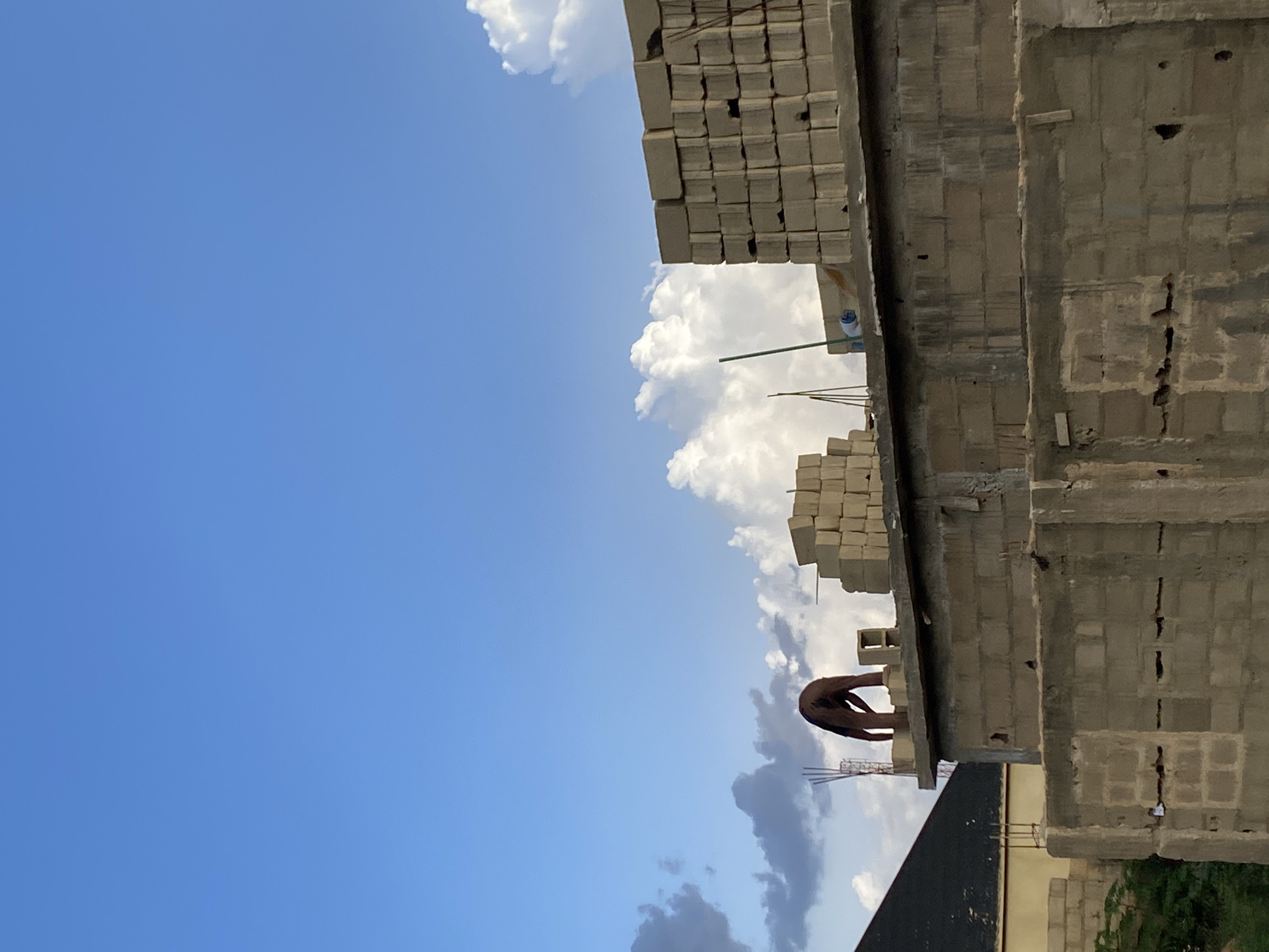 A person is working on a construction site. The scene is outdoors, with a clear blue sky and some white clouds. A partially constructed building made of concrete blocks is visible. Stacks of concrete blocks are also present on the site. The person, wearing dark clothing, is bent over, seemingly engaged in some task related to the construction. The angle of the image is unusual, with the ground appearing to be on the left side of the frame and the sky on the right. This suggests the photo may have been taken from an elevated position or that the camera was rotated. The lighting indicates it is daytime, likely morning or afternoon, given the brightness of the sky. The overall atmosphere is one of ongoing construction work.
