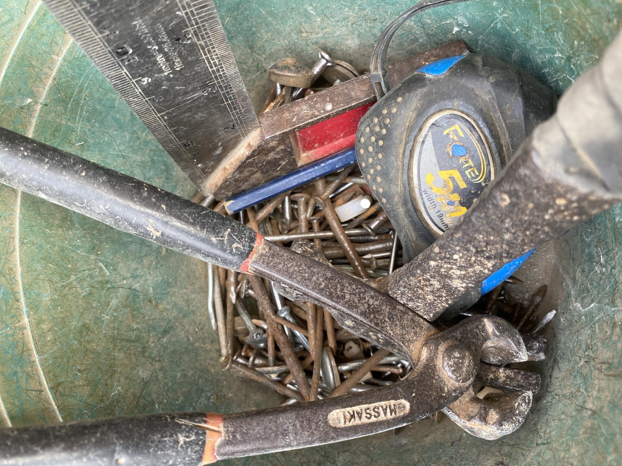 In a green plastic tub, a collection of rusty nails is visible. Among the nails are a pair of bolt cutters, a tape measure, and a metal ruler. The bolt cutters have black handles with orange accents. The tape measure is black and round with a yellow and blue label that reads "FORTE." The metal ruler is angled and marked with measurements. A small red and blue object is partially visible behind the ruler. The scene appears to be a close-up, overhead shot of tools and supplies.