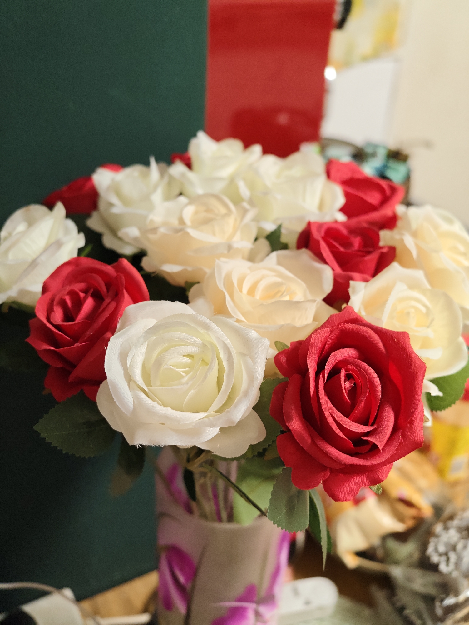 A close-up, eye-level, medium shot captures a bouquet of artificial roses in various shades of red, white, and cream, arranged in a patterned vase. The roses, with their velvety petals, are densely packed, with some fully bloomed and others still in bud. Dark green leaves peek out from between the flowers, adding a natural contrast to the artificial blooms.
The background is softly blurred, suggesting an indoor setting. To the left, a dark green surface dominates, possibly a wall or fabric backdrop. To the right, out-of-focus elements suggest a shelf or surface with various objects, including hints of red and yellow.
The lighting appears to be artificial, casting subtle shadows and highlighting the texture of the rose petals. The overall impression is one of a decorative arrangement, likely placed on a table or shelf. No specific landmarks or features identifiable with Petah Tikva, Israel are present in the image.