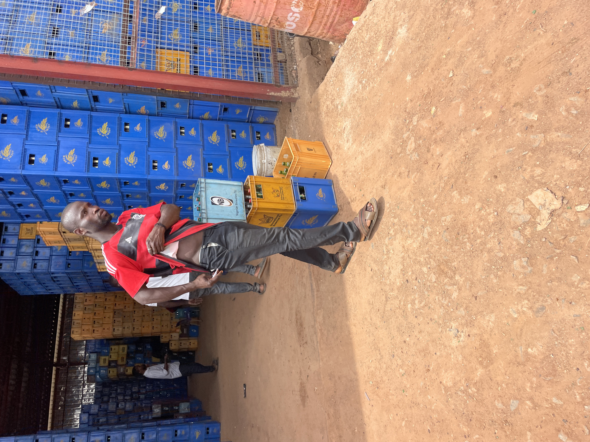 Outside a warehouse in Gura Top, Nigeria, a man wearing a red and black shirt, dark pants, and sandals stands near stacks of blue and yellow crates. Another man in a white shirt is further back. The ground is reddish-brown dirt. An orange barrel is visible in the background.