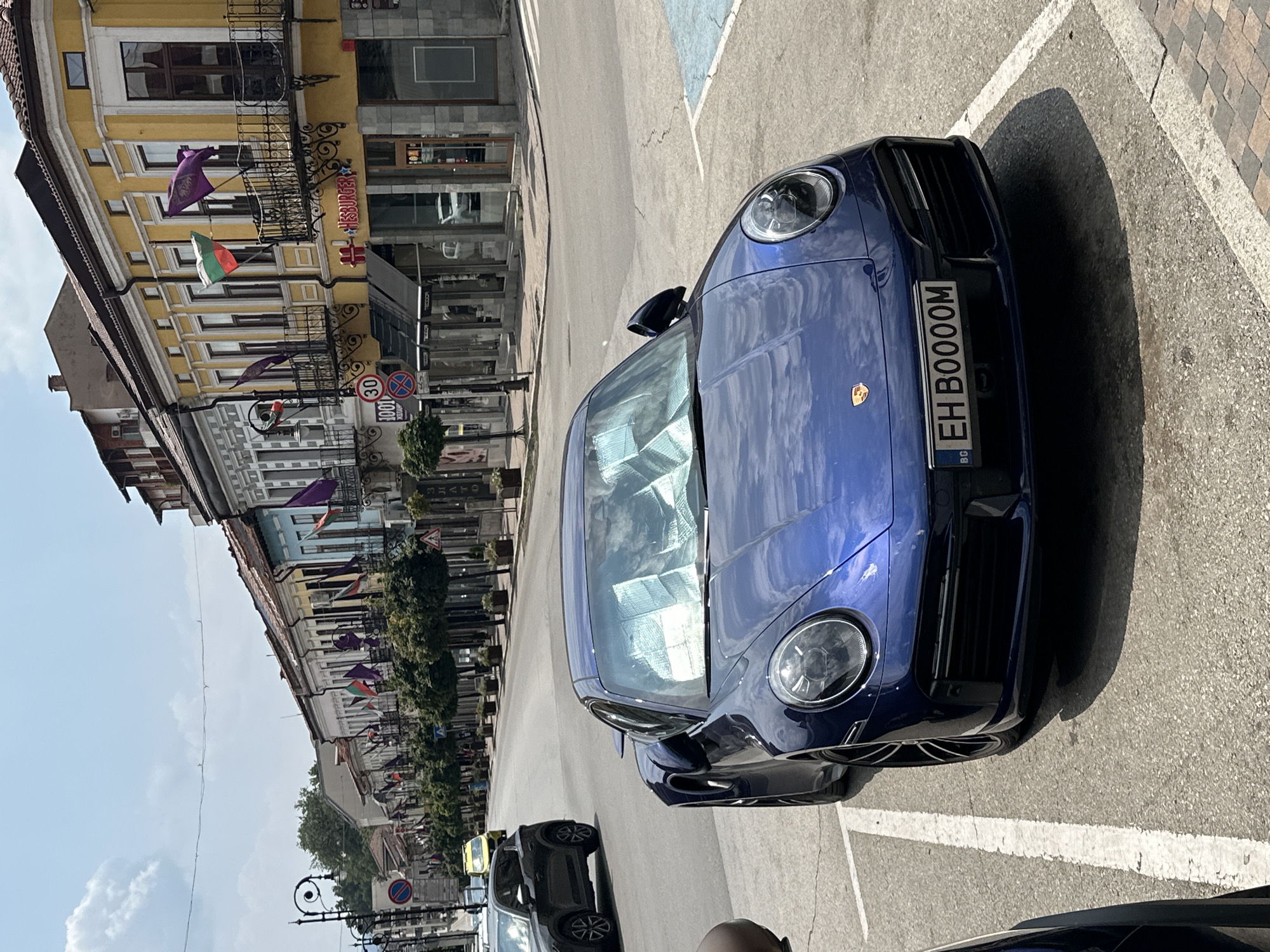 The image shows a dark blue Porsche 911 Turbo S parked on a street in Veliko Tarnovo, Bulgaria. The car has a custom license plate that reads "EH BOOOOM". The street is lined with buildings, some of which are decorated with flags. In the background, a yellow taxi and a black SUV are visible. The sky is clear and the sun is shining. The street appears to be a commercial area with shops and restaurants. The buildings are in a traditional European style with colorful facades and ornate balconies. The scene is set in the old town of Veliko Tarnovo, likely on Tsar Ivan Asen II Boulevard, a prominent street in the city known for its historic architecture and vibrant atmosphere.