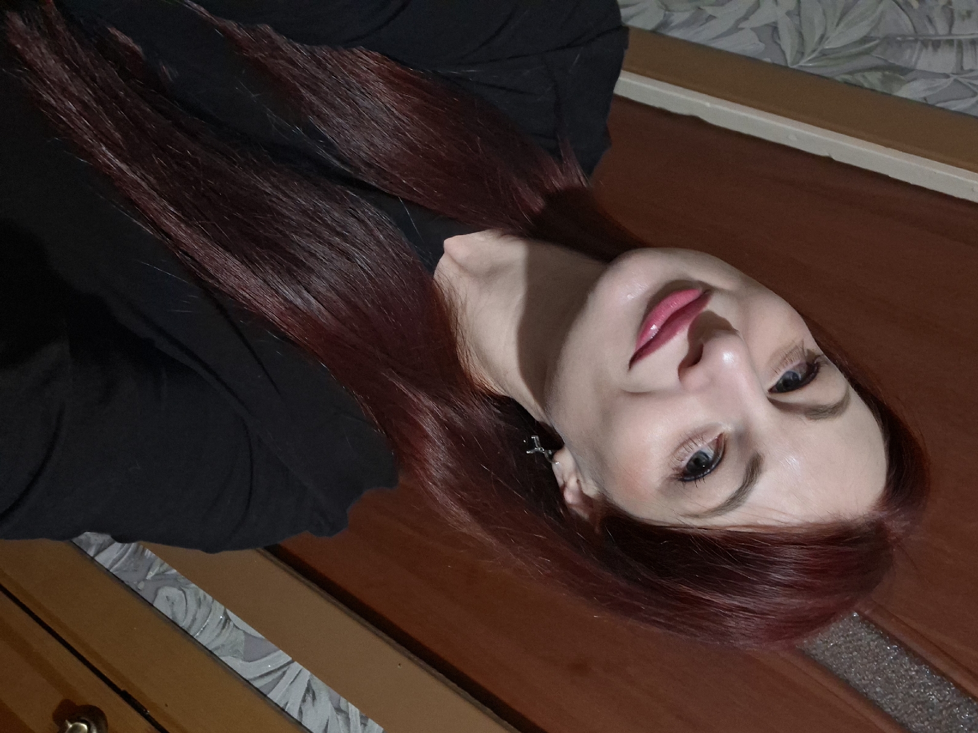 A woman is pictured indoors. She has long, auburn hair and is wearing a black top. She is also wearing an earring. The background features a wooden surface.