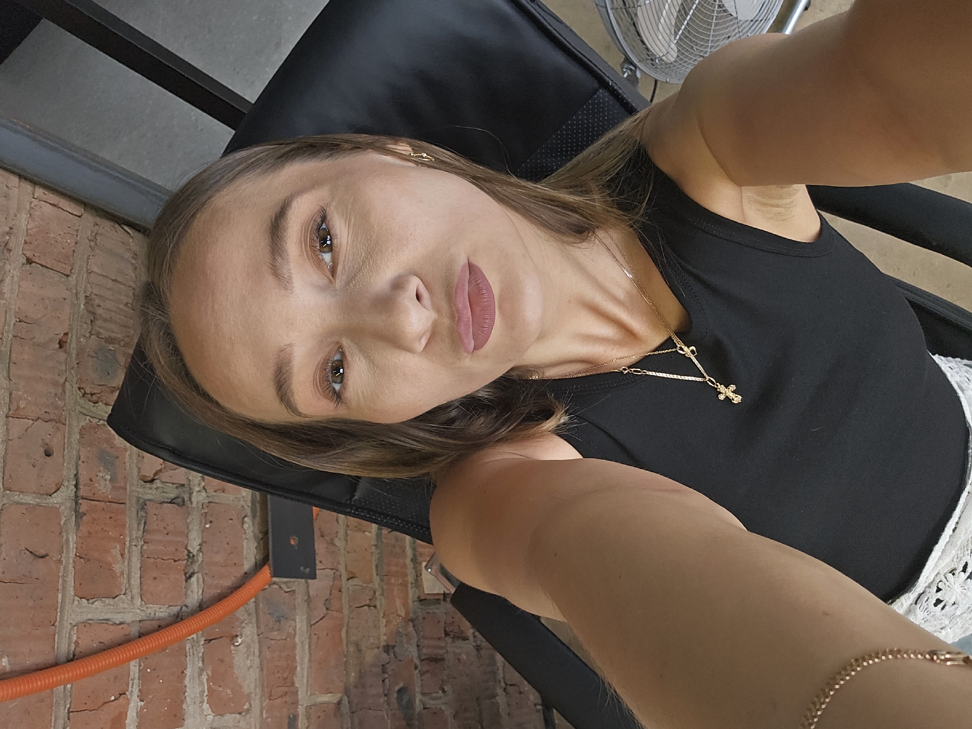 The image is a selfie of a woman with fair skin, light brown hair, and light brown eyes, wearing a black tank top and a gold necklace with a cross pendant. She is sitting in a black chair against a brick wall and is positioned at an angle. An orange pipe runs along the base of the brick wall. A fan can be seen in the background. The location is not specified, but the prompt mentions Chaikovsky, Russia.