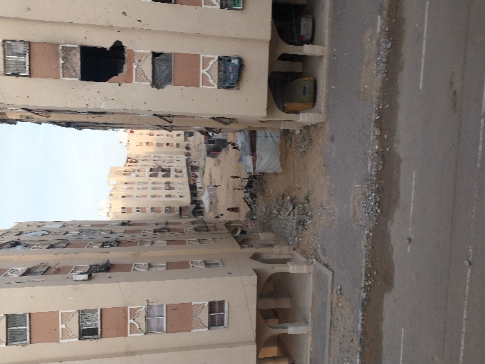 This image presents an urban landscape in Khan Yunis, Palestinian Territory, observed from an elevated vantage point overlooking a street and an inner block area. In the foreground, a paved road with two faded white lane markings is visible, its edge strewn with small rocks and dirt.
Two multi-story residential buildings, light-beige with rust-colored vertical strips and white decorative window accents, frame the central view. The building on the right exhibits significant structural damage, including a large, irregular dark cavity on an upper floor, shattered windows, and numerous impact marks on its facade. Below, its ground floor features open arches where a large, tarp-covered vehicle and a white sedan are parked. The building on the left also displays signs of wear and damage, with some windows boarded or broken.
Between these two primary structures, an open expanse of sandy earth is heavily covered with piles of rubble, broken concrete, and twisted metal debris. Scattered within this area are several rudimentary shelters constructed from tarpaulins and corrugated sheets. Multiple individuals, primarily appearing as children, are dispersed across this ground, some walking; one person is positioned near a bicycle. Further in the background, additional multi-story buildings of similar architectural style complete the urban setting under a clear sky.