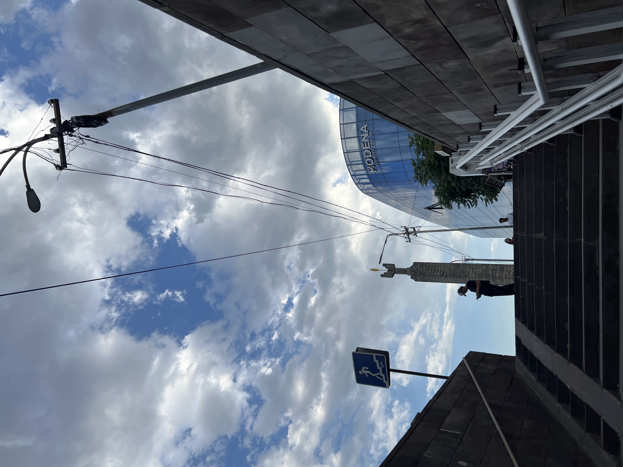 The image depicts an outdoor scene with a cloudy sky. In the foreground, a set of dark-colored stairs ascends along the right side of the frame. Above the stairs, a modern, curved building with a glass facade is visible, bearing the text "MODENA BUSINESS CENTER." A utility pole with multiple wires and a street lamp is also present, extending across the sky. In the middle ground, a tall, slender monument is visible, and a sign with a blue background shows a white silhouette of a person climbing stairs, indicating a pedestrian pathway or stairs. A person in dark clothing is standing on a lower platform, looking at their phone. The overall scene suggests an urban environment, possibly in Yerevan, Armenia, given the provided location context. The time of day appears to be daytime, with scattered clouds and patches of blue sky suggesting variable weather conditions. The presence of a business center and a monument points to a public or commercial area.