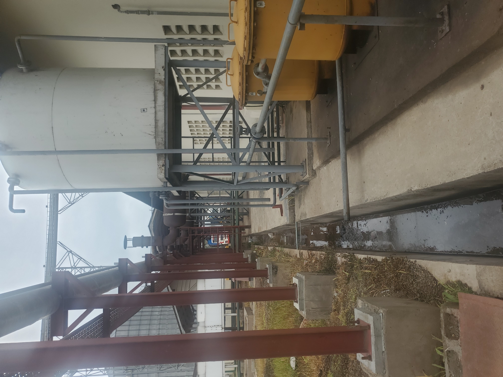 The image captures an outdoor industrial scene in Gwagwada, Nigeria. Dominating the foreground are large, industrial pipes supported by reddish-brown metal beams. To the right, two bright yellow cylindrical tanks with domed tops and handles are visible. These tanks are connected by piping. Further back, a tall white cylindrical tank rests on a metal platform. Adjacent to the building, a series of rectangular vents are present. A concrete drainage ditch runs along the ground, filled with dark, murky water. The ground is a mix of concrete and dry grass. In the background, the skeletal framework of an elevated structure and parts of large, silver grain silos can be seen. A truck with a red cabin is partially visible in the distance, behind the pipes. The sky is overcast.