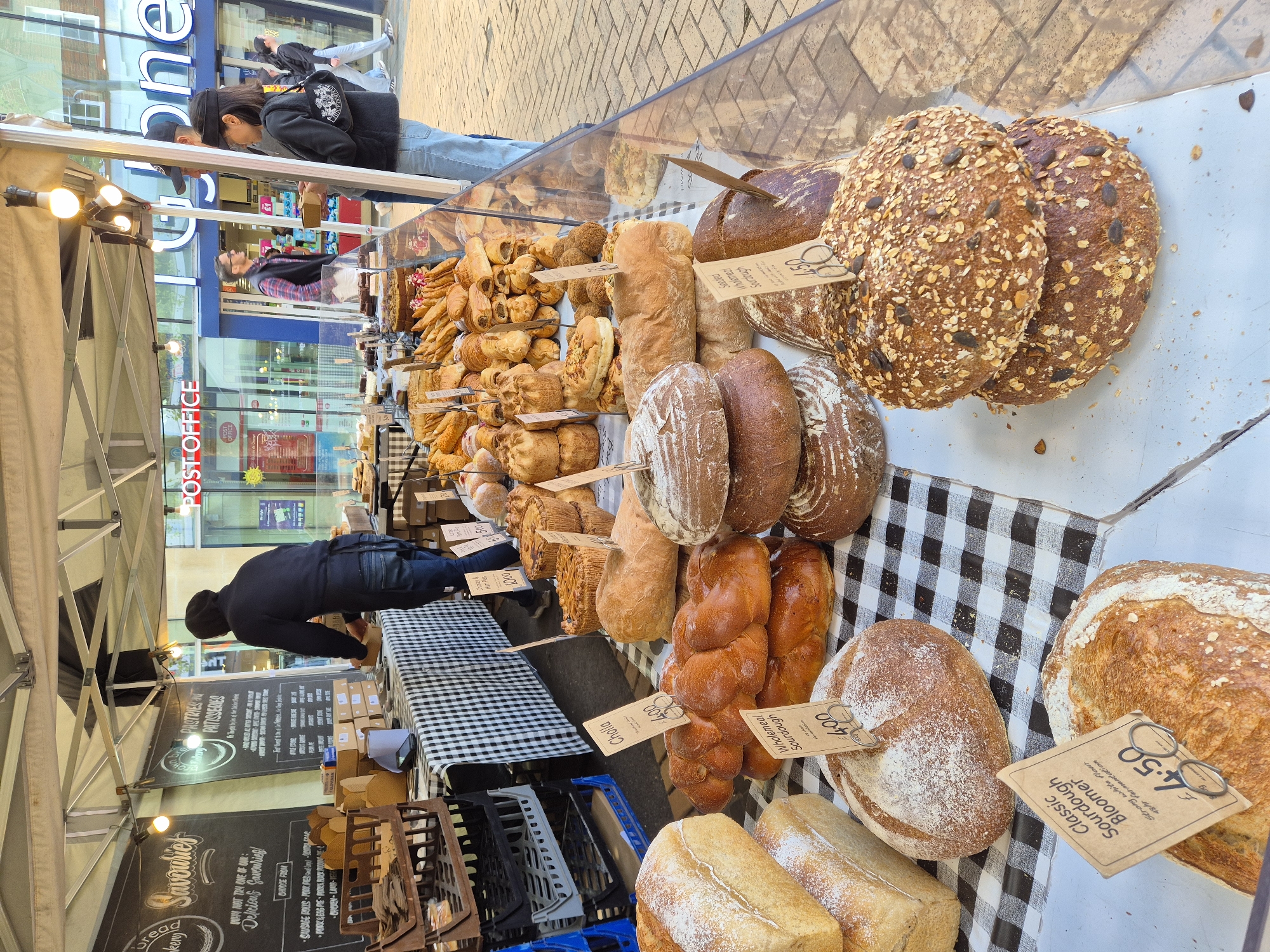 The image shows a bread stand in Chelmsford, United Kingdom, near a "Post Office" and "Fone" store. The stand has a variety of bread loaves and pastries displayed on a checkered tablecloth. A man is reaching over the display to purchase bread, and a couple is standing near the glass display. A sign lists various "Savouries" and "Pastries". The bread prices are displayed on small tags. The stand is set up under a tent-like structure with overhead lights. Crate holders sit underneath the table.