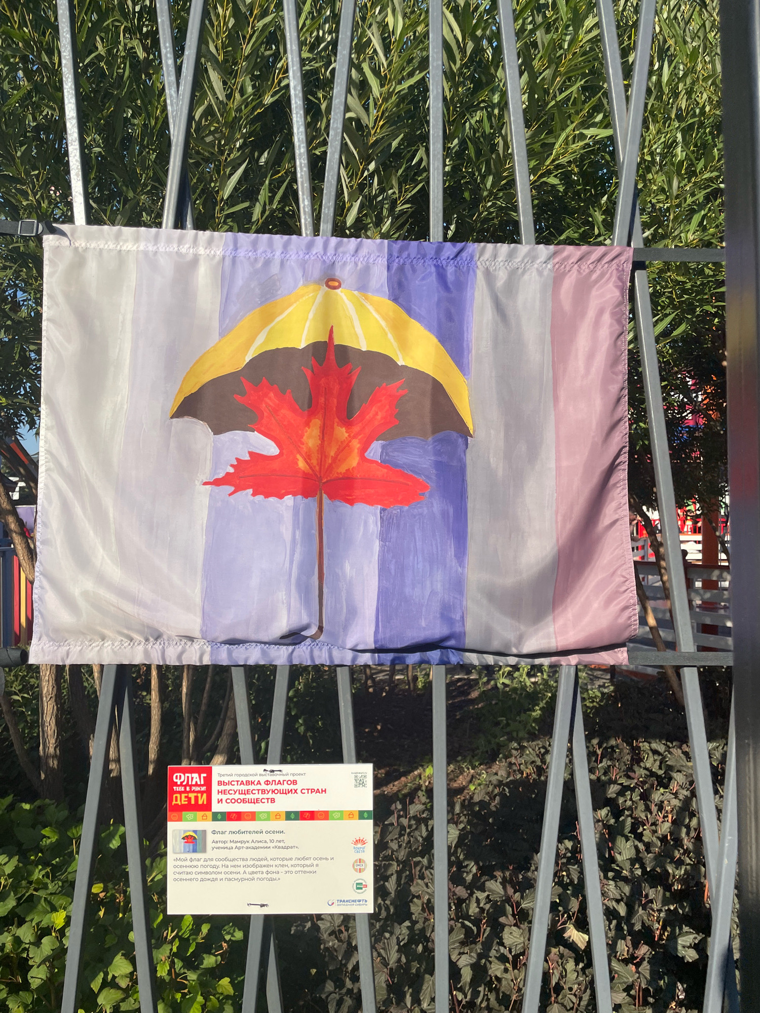 A flag depicting a yellow umbrella over a red maple leaf is displayed on a fence in Omsk, Russia. The flag is attached to a metal fence with green foliage in the background. A sign beneath the flag provides details about the artwork, including the artist's name, age, and a description of the flag's symbolism. The sign also indicates that the flag is part of the "Flags of Non-Existent Countries" exhibition. The exhibition is part of a city-wide project, and the sign features the logo of "Transneft," an energy company that sponsors the project. The weather appears to be sunny, with shadows cast on the fence and ground. The scene suggests an outdoor public art installation.