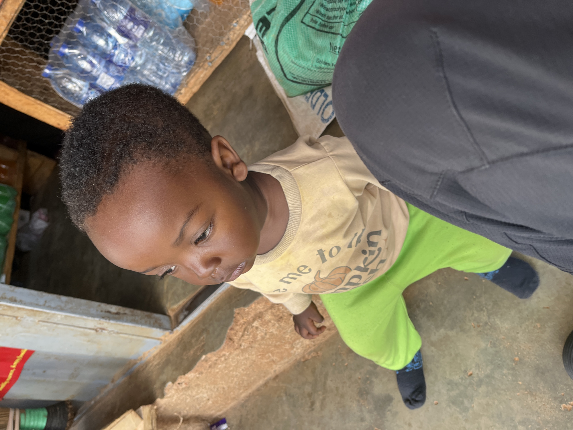 A young child is sitting on a concrete floor in an outdoor market setting in Barakin Akawo, Nigeria. The child is wearing a light yellow long-sleeved shirt with a pumpkin graphic and the words "me to" and "kin" visible, along with bright green pants and dark socks. The child is looking to the right of the frame. Behind the child, stacked water bottles are visible in a wooden crate with wire mesh. A large green bag with text and a white bag with blue text are also present on the ground. To the left, a weathered doorway and a rough, textured surface are visible. The ground is littered with small debris.