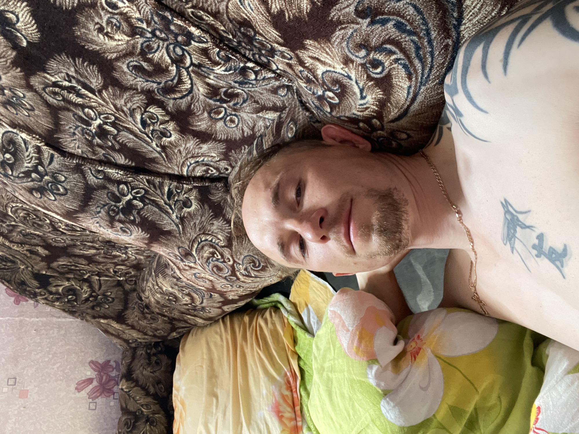 A male individual is lying on a bed in a room. He has light skin, short blond hair, a goatee, and is wearing a gold chain. He has tattoos on his upper left arm and chest. The bed is covered with a patterned brown fabric, and there are pillows with floral patterns. The background shows a wall with a floral wallpaper. The scene is shot from a high-angle perspective. The location is identified as Nizhnii Bugrysh, Russia.