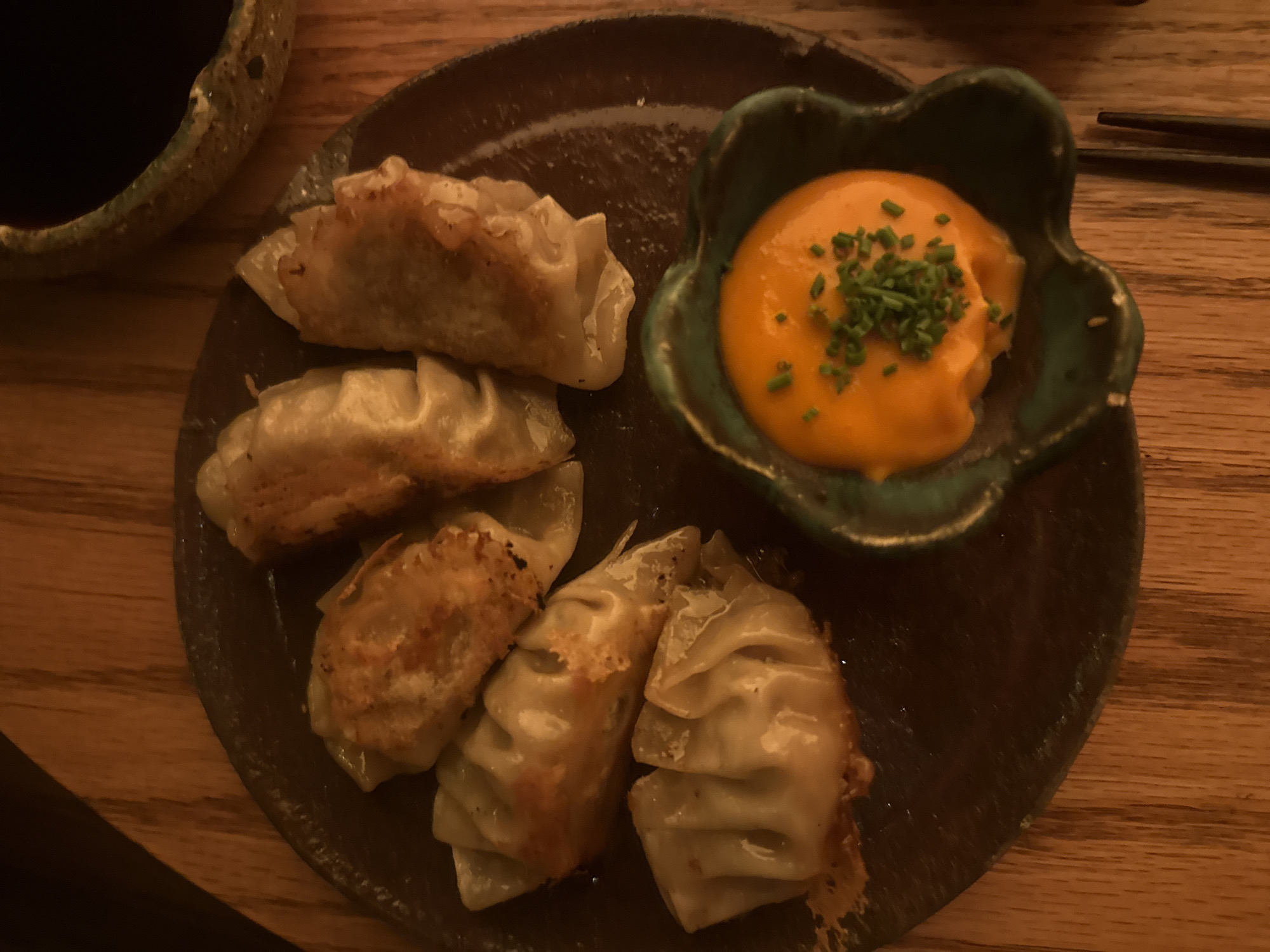 The image shows a plate of dumplings and a small dish of orange sauce with green herbs, likely chives, on a wooden surface. The dumplings appear to be pan-fried, with a slightly crispy exterior. A small bowl filled with dark liquid (possibly soy sauce) is visible in the background. The scene is likely a restaurant setting. There is no evidence to determine if the image was taken in Mexico City.
