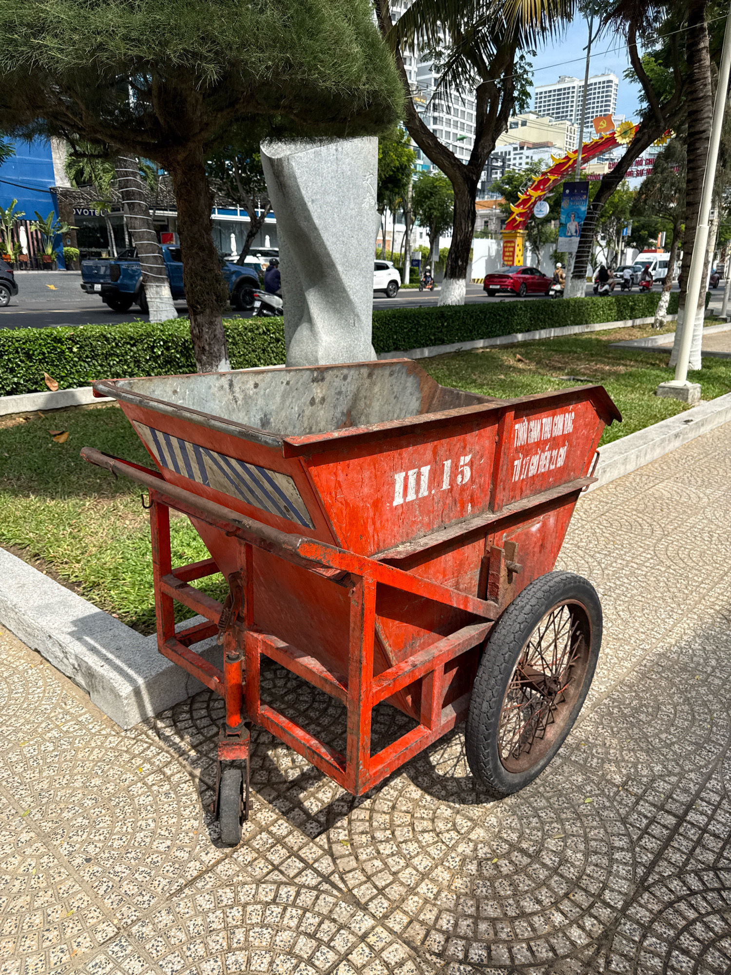 This is a medium-shot, eye-level photograph of an old, weathered red metal pushcart, likely used for trash collection, parked on a patterned tile sidewalk in Nha Trang, Vietnam. The cart is in the foreground, taking up a significant portion of the frame, and is slightly to the left of center. It has a large, rectangular open bin at the top, with visible rust and grime. On the side of the bin, there are white painted markings: "III.15" in large numerals and a line of text in Vietnamese, which appears to be an instruction or identification related to garbage collection.
The cart has two large spoked wheels at the rear and a single smaller, fixed caster wheel at the front. The metal frame supporting the bin is a simple, robust construction. A thick, rusty metal bar extends from the left side of the cart, serving as a handle for pushing.
Behind the cart, a green grassy area borders the sidewalk, separated by a low concrete curb. Beyond the grass, a street is visible with light traffic, including a blue pickup truck, a red car, and several motorbikes with riders. Trees with dark bark and lush green foliage are scattered throughout the scene. In the background, modern buildings with many windows rise into the sky, typical of a city environment. One building prominently displays a blue facade with the word "NOVOTEL" visible. A festive red and yellow dragon archway or decoration spans across the street in the middle distance, possibly indicating a celebration or cultural event. Palm trees are also present, adding to the tropical feel of the location. The lighting suggests it is daytime, with shadows cast by the cart and surrounding vegetation.