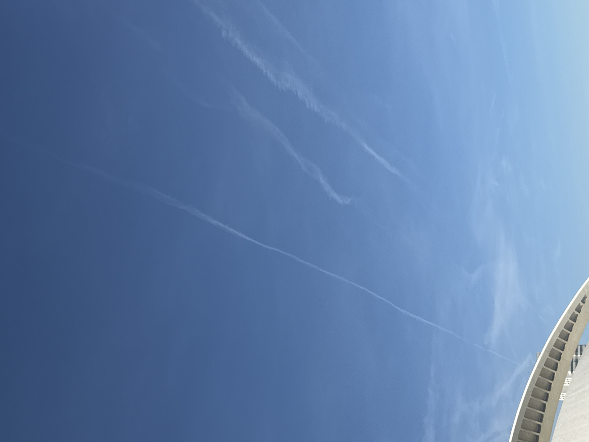 The image shows a clear blue sky with several contrails, likely from airplanes. In the lower right corner, part of a white, arched architectural structure is visible, which is likely the Pont de l'Assut de l'Or, a bridge in Valencia, Spain designed by Santiago Calatrava. There are no people visible.