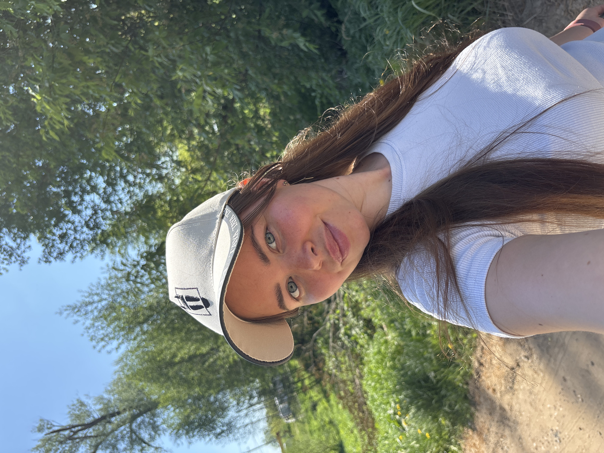 A young woman is taking a selfie. She is wearing a white t-shirt and a light-colored baseball cap. She has long brown hair and green eyes. The background suggests an outdoor setting, possibly near a dirt path or road, with greenery and trees in the distance. Based on the location data provided, the location may be Malyi Bukor, Russia. The image's angle suggests it was taken with the phone above her.