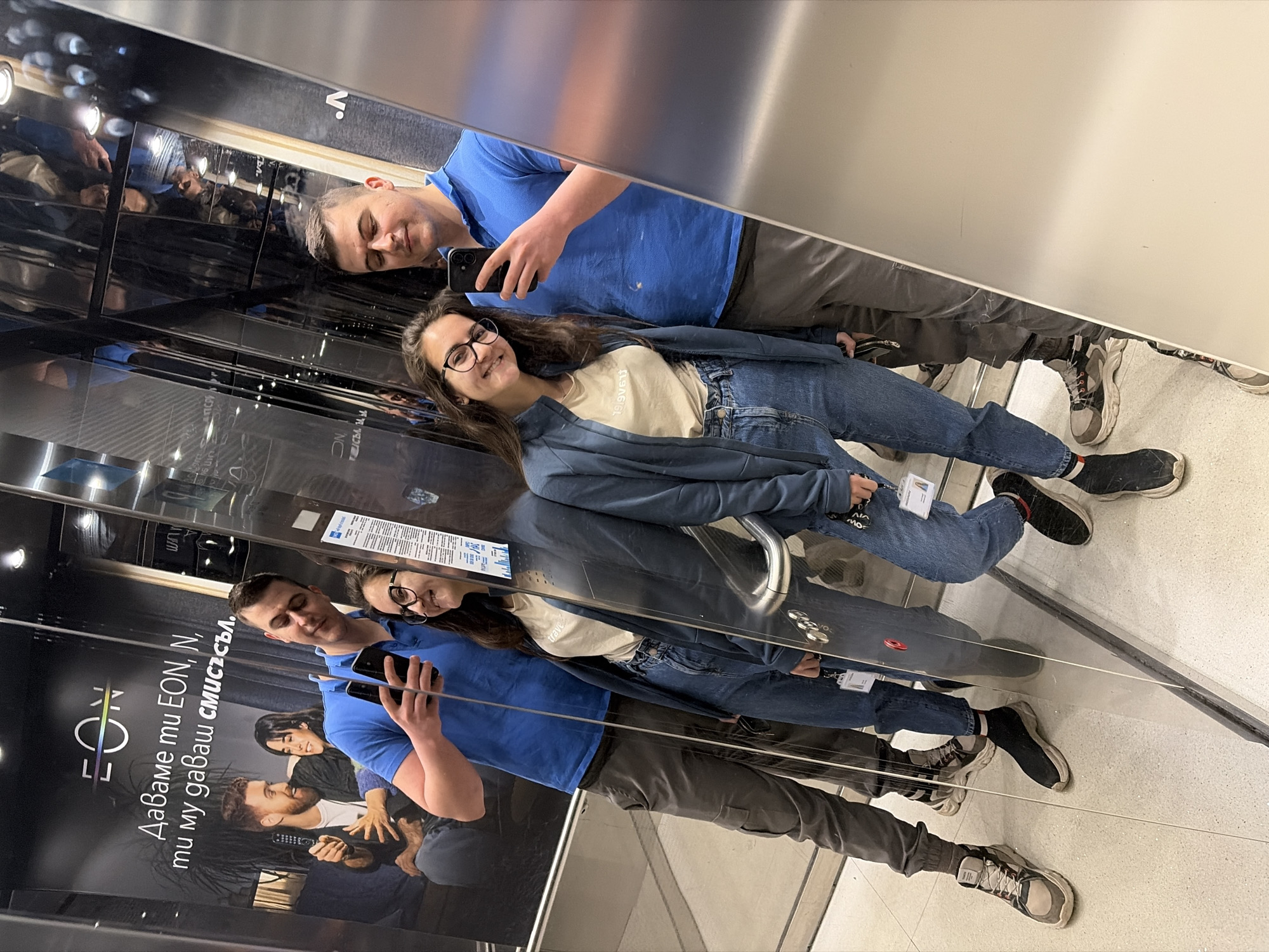 The image shows a man and woman standing in an elevator with mirrored walls. The man wears a blue t-shirt and gray pants, holding a smartphone to take a picture in the mirror. The woman has long brown hair, glasses, a beige t-shirt, a blue jacket, and blue jeans. A poster for EON, written in Bulgarian, is visible on the wall, which mentions Sofia, Bulgaria.