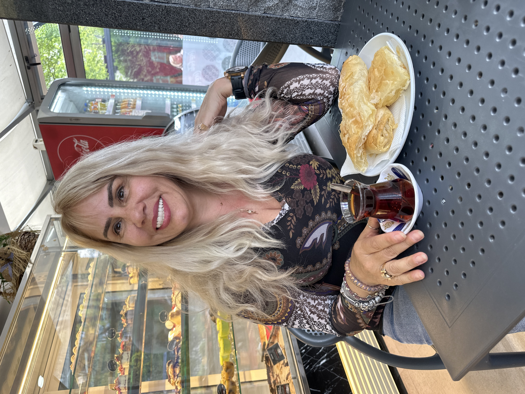 The image captures a cheerful woman seated at an outdoor or semi-outdoor cafe in Sofia, Bulgaria, enjoying a tea and pastry break. The photo is rotated 90 degrees counter-clockwise.
The primary subject is a smiling woman with long, voluminous blonde hair, looking directly at the camera. She wears a dark, long-sleeved top adorned with floral and paisley-like patterns. Her left wrist displays a watch and several bracelets, while rings adorn her fingers.
In front of her, on a dark gray, perforated table, sits a white plate holding three golden-brown pastries, likely a type of börek, resting on a white paper napkin. Beside the plate is a traditional tulip-shaped Turkish tea glass filled with dark tea, placed in a white saucer with blue and white decorative patterns.
The background reveals elements typical of a cafe setting. To the left of the woman (if oriented correctly) is a red "Coca-Cola" branded refrigerator, visible through glass, containing various bottled beverages. Further back, a glass display case showcases an assortment of pastries or desserts. Green foliage can be seen through a window or open area, suggesting a pleasant daytime setting. The woman appears relaxed and happy, indicated by her warm smile.