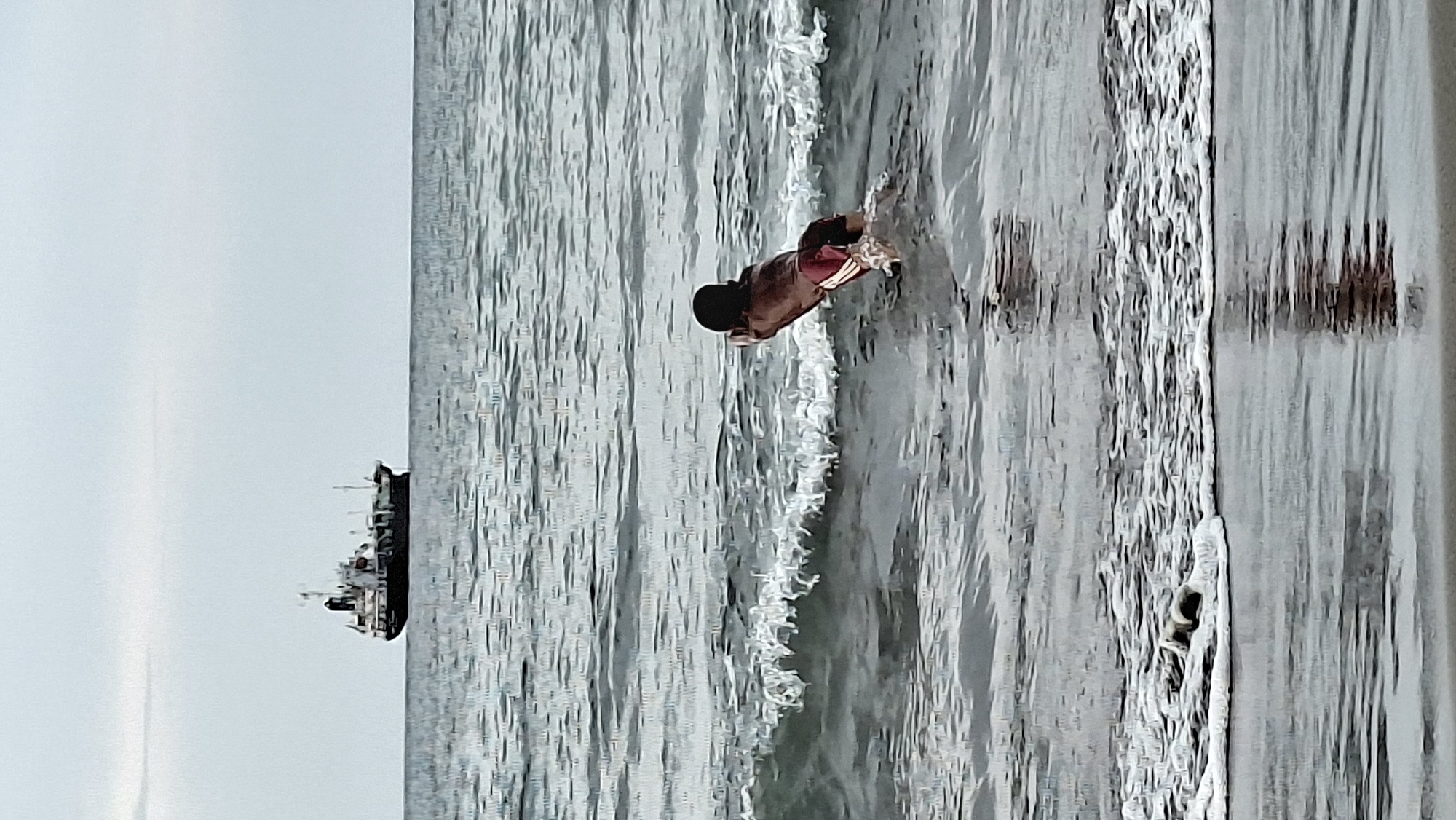 A person is swimming in the ocean near the coast of Lhokseumawe, Indonesia. The person is wearing red and white striped shorts and is partially submerged in the water. A cargo ship is visible in the distance on the horizon. The water is choppy with small waves. The sky is overcast. The image is taken from a low angle, looking out towards the ocean.