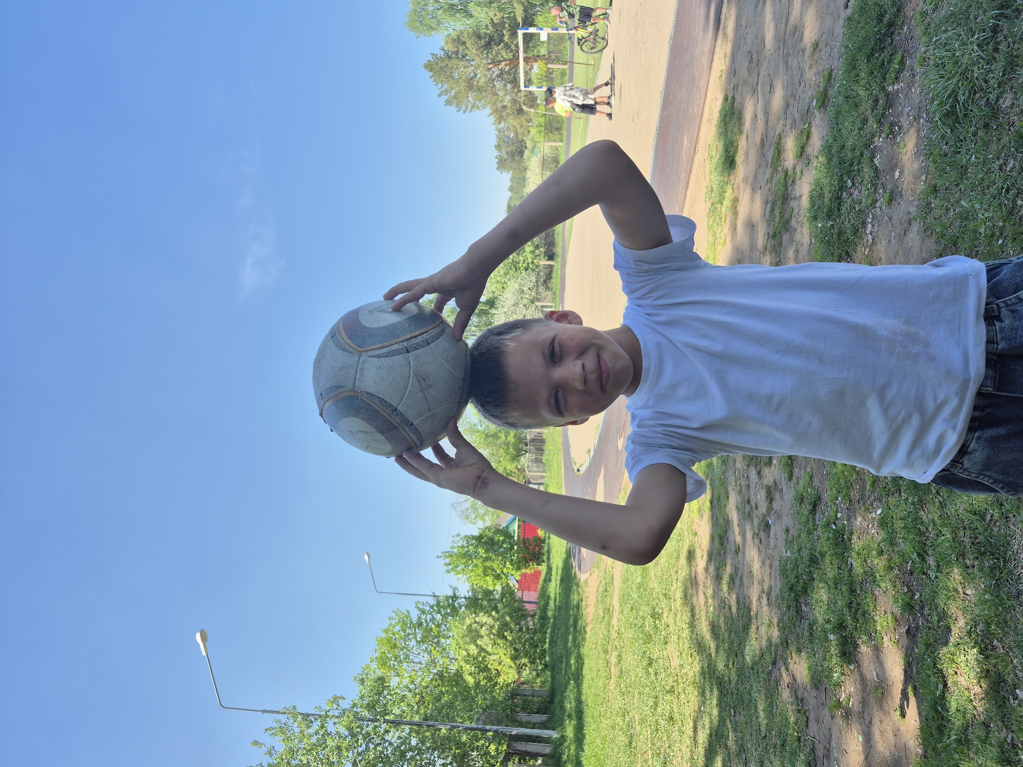 A boy is holding a soccer ball over his head outdoors in the city of Chaikovsky, Russia. He is wearing a white t-shirt and jeans. The background shows grass, trees, a blue sky, a light pole, and other people in the distance near a soccer goal.