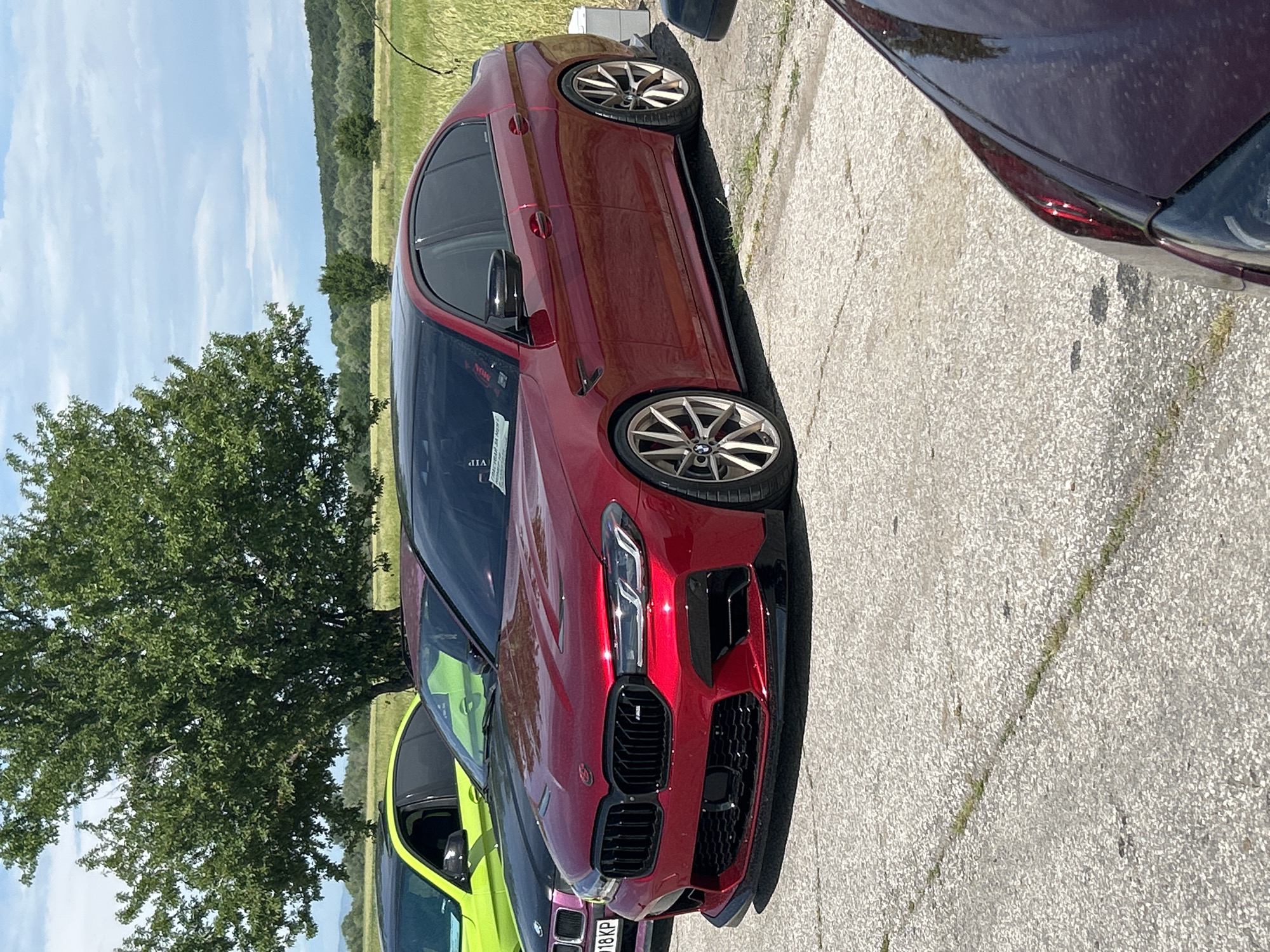 A maroon BMW M5, parked on a concrete surface, is the central subject of the image. The car is positioned next to a green vehicle and a dark-colored car, both partially visible. A tree stands to the left of the BMW. In the background, a field is visible under a partly cloudy sky. The location is Dalbok Izvor, Bulgaria.