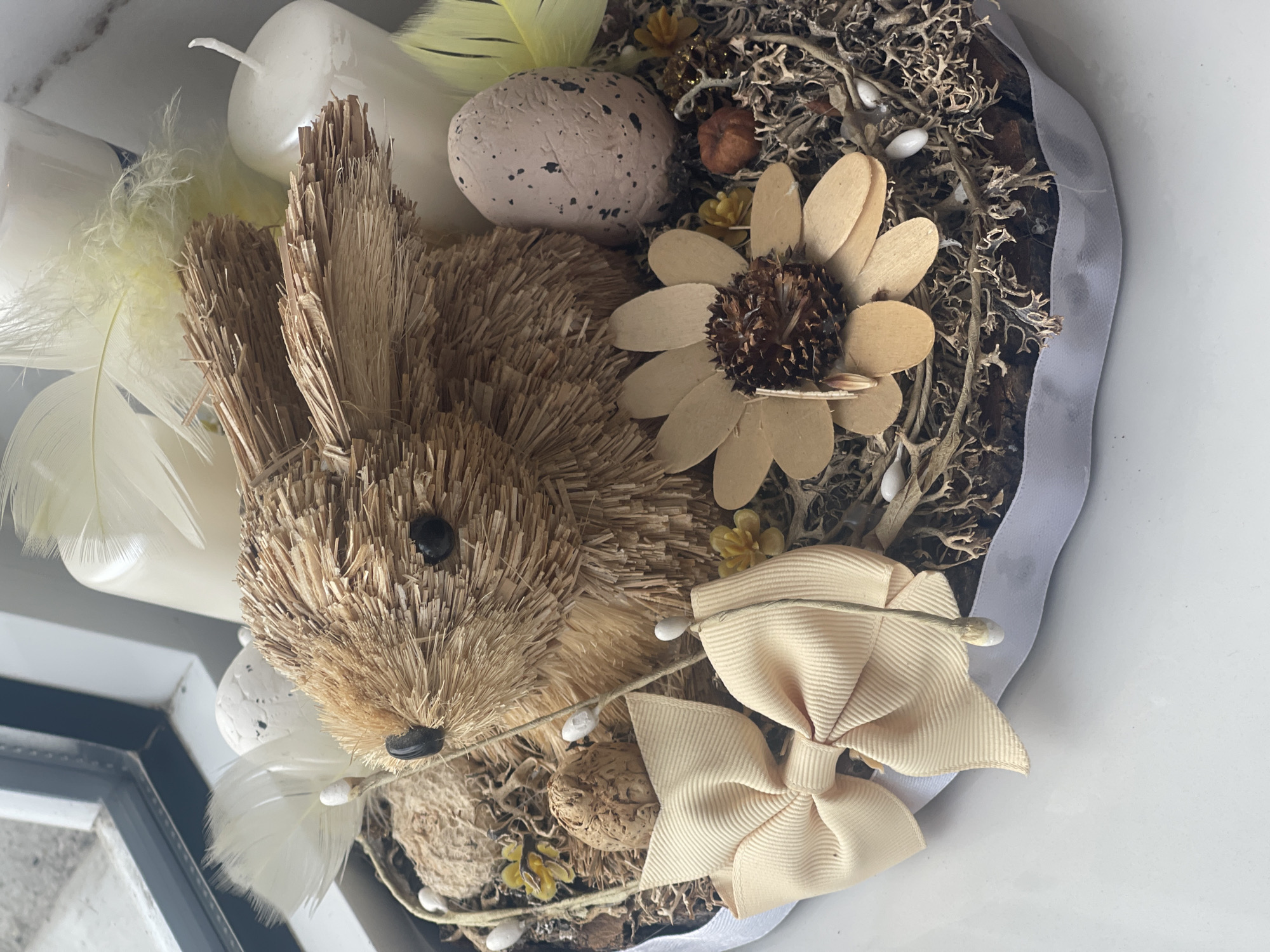 This is a close-up, top-down view of an Easter-themed decorative arrangement. The central subject is a straw-like rabbit figurine, positioned slightly left of center. It has black bead eyes and a black seed for a nose. Surrounding the rabbit are various decorative elements: a speckled egg, white feathers, a wooden flower with a brown center, and a light beige ribbon tied into a bow.
The arrangement is set within a dark, textured base that appears to be composed of moss and bark. There are also several white candles, one of which is partially visible behind the rabbit's head, and small, white, egg-shaped accents are scattered throughout. A light blue and white ribbon borders the arrangement on the right side.
The lighting suggests it is daytime, possibly indoors, as there are no strong shadows indicative of direct sunlight. The overall mood is festive and decorative, likely intended for an Easter celebration. There is no visible text in the image.