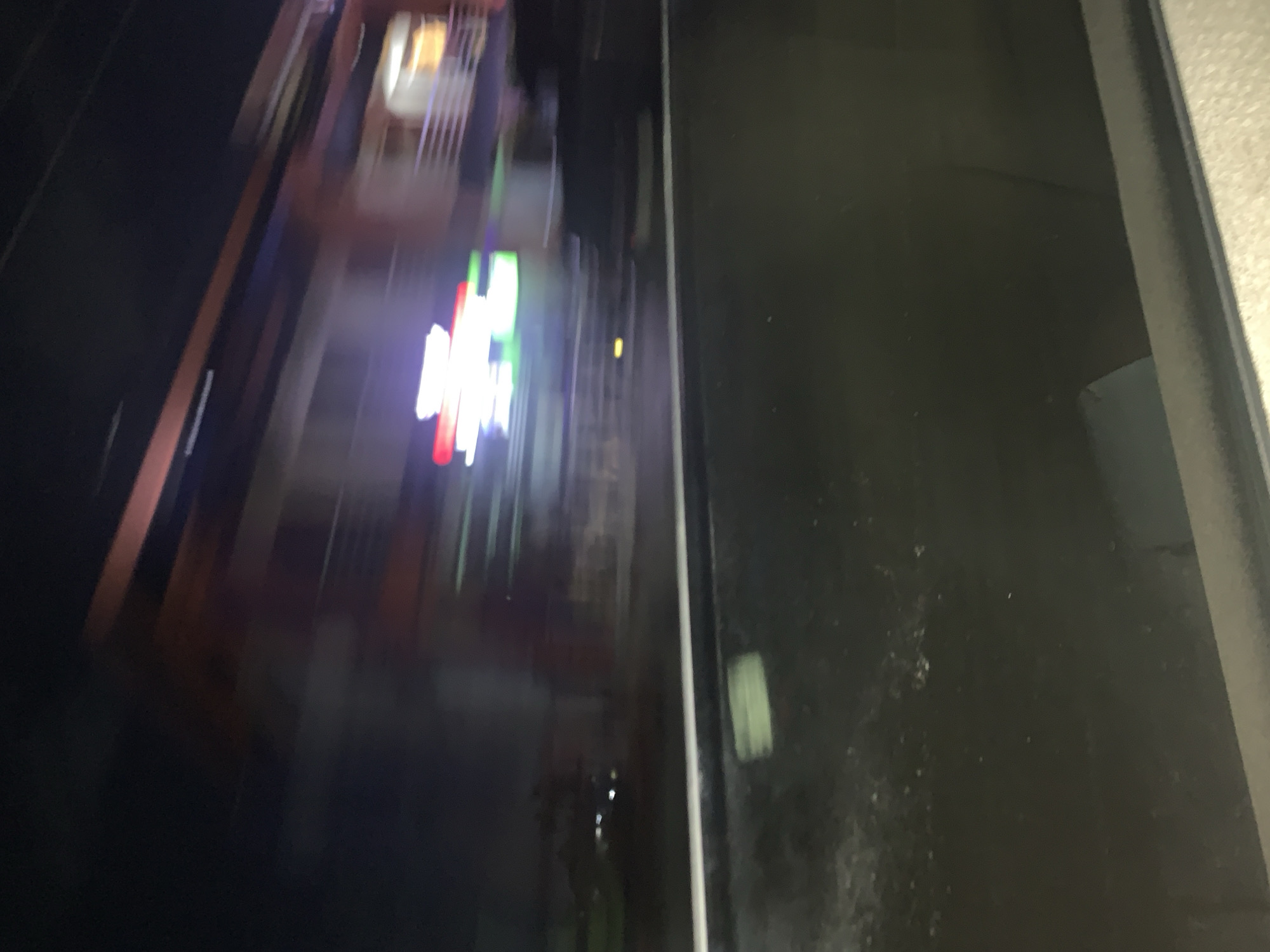 The image is an abstract, blurred view of a city at night. Visible are illuminated buildings with multi-story architecture and bright neon signs. The buildings appear to be located along a road or street, evident by the vertical orientation of the scene. The right portion of the image shows a dark, speckled surface with a slight gradient, possibly part of the camera lens or a dirty window. The exact location in Dar es-Salaam, Tanzania, is not identifiable from the blurred details of this image.