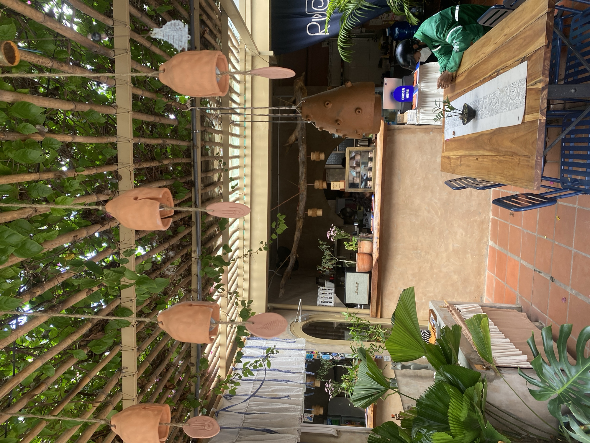 The image depicts an outdoor seating area of a cafe in Mũi Né, Vietnam. A wooden table with blue metal chairs is set in the foreground, with a small potted plant on a decorative runner. A person wearing a green uniform, helmet, and mask is seated at the table, looking down at their hands. The background features a counter area with a Marshall speaker, potted plants, and a blue curtain with white lettering that reads "RUCA". Above, a pergola structure is covered with lush green foliage. Several terracotta-colored bells hang from the pergola, adding to the rustic ambiance. The floor is paved with terracotta tiles.