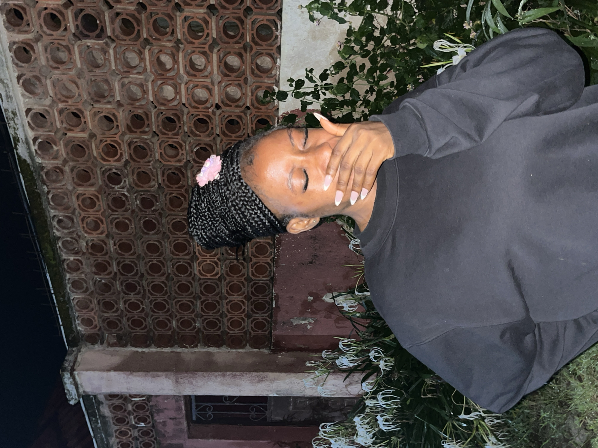 A medium shot captures a Black woman with braided hair, adorned with a pink flower, covering her mouth with her right hand. Her eyes are closed, and her expression suggests laughter or amusement. She is wearing a black crewneck sweatshirt. The background features a wall with a distinct brick pattern of concentric circles, a weathered pink pillar, and lush white spider lilies. The overall scene evokes an outdoor setting at night, possibly in Jos, Nigeria, given the architectural style and flora.