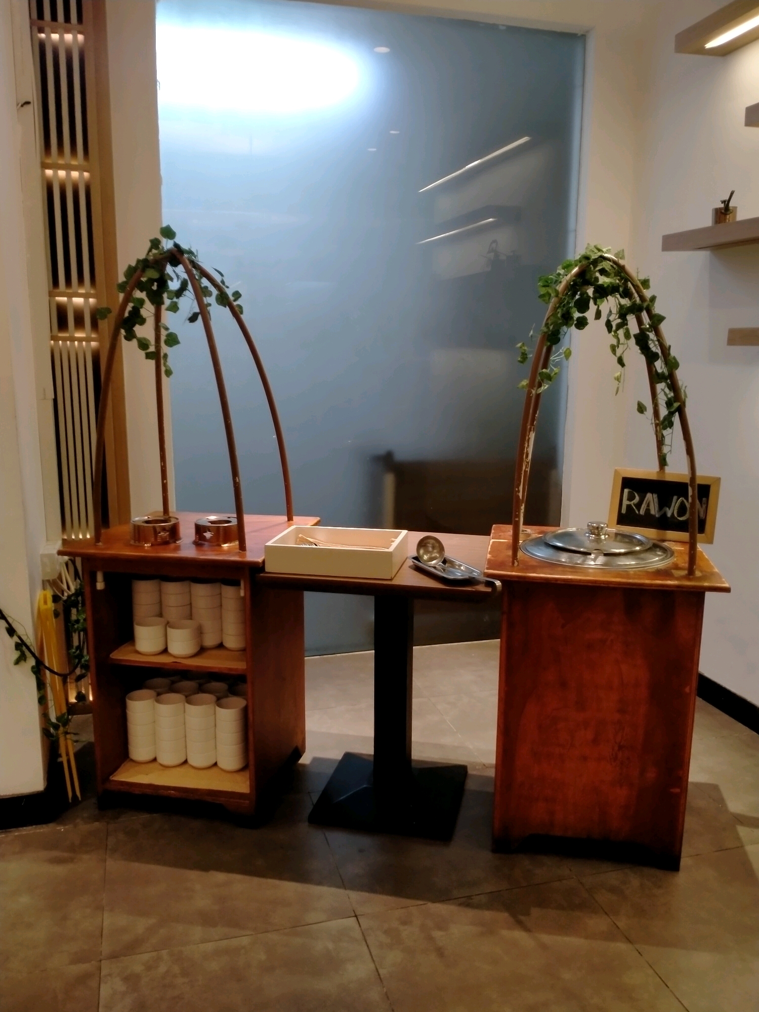 This is a medium shot, eye-level, outdoor photograph of a food serving station at the Jatim Park 2 food court in Batu, Indonesia. The station is made of dark wood and is decorated with artificial ivy. It features two serving counters, each with a large metal pot for food. A chalkboard sign on the right counter reads "RAWON." The left counter holds stacked white cups and bowls. The station is on a tiled floor in front of a large, frosted glass window.