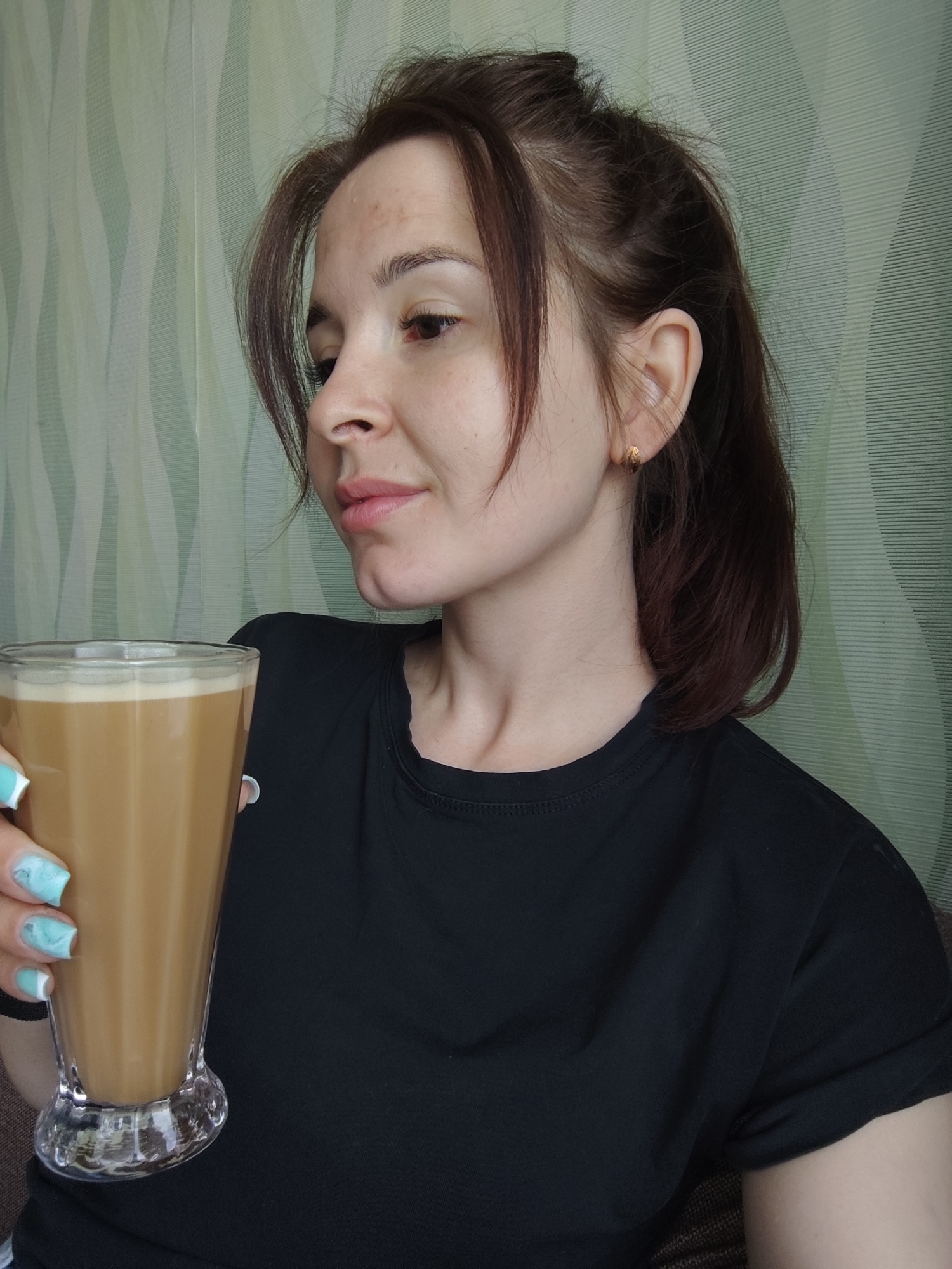 A woman with brown hair in a ponytail and a black t-shirt is holding a glass of latte. She is looking to the left, and her nails are painted with a light blue and white marble design. The background is a light green patterned wall. It is not possible to verify the exact location within Фоки, Russia, from this image.