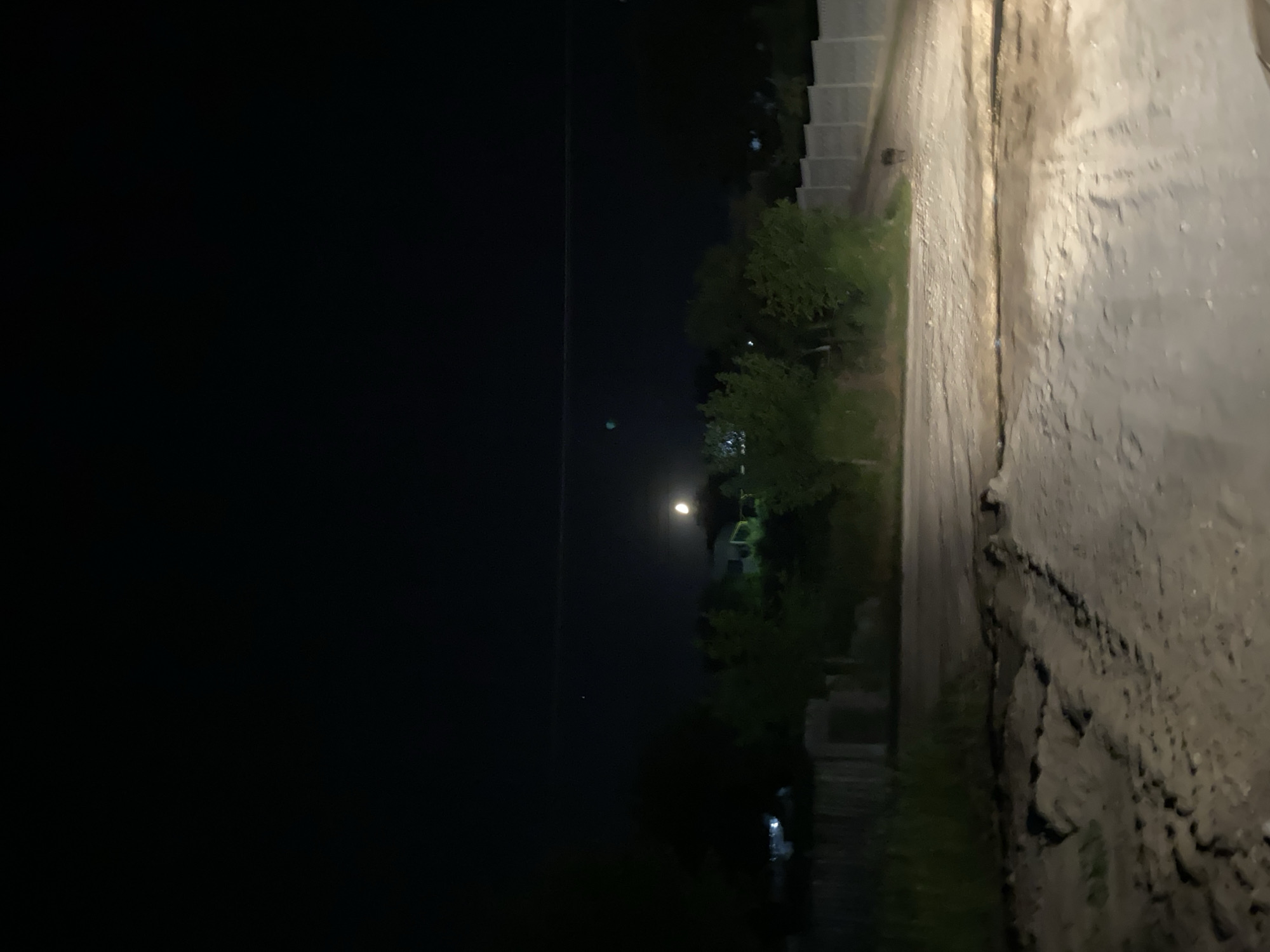 The image captures a nighttime scene in Negrești, Moldova. The perspective is from a ground-level view, looking across a narrow, rough dirt path. To the right of the path, a textured, uneven embankment rises, appearing to be made of packed earth or gravel with visible erosion and debris. The path itself is a lighter color, suggesting it's made of dirt or fine gravel.
To the left of the path, a dark, open expanse dominates the left side of the frame, suggesting a void or very dark area, possibly a field or undeveloped land. In the middle distance, beyond the path, several trees with dense foliage are visible, illuminated by artificial lights. These lights create a faint glow in the darkness. Further in the distance, faint outlines of buildings or structures can be discerned, some with hints of lights. One particular light source in the distance appears to be a streetlamp or a bright overhead light, casting a visible beam into the dark sky. The overall atmosphere is dark and quiet, typical of a rural nighttime setting. There is no visible text in the image. The time of day is clearly night, and no specific weather conditions are apparent.
