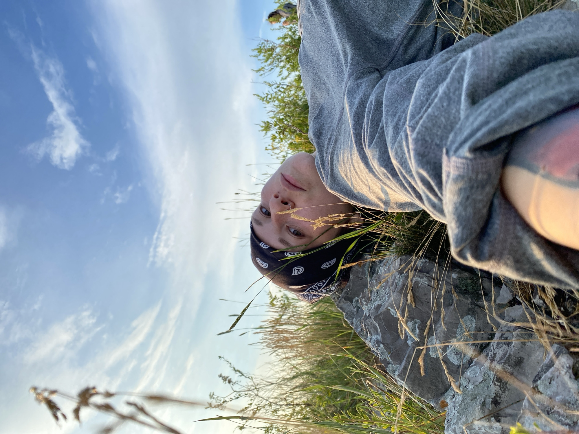 In Kondurovka, Russia, a person lies on their back, resting their head on a grey, textured rock. They wear a black bandana with a white paisley pattern and a grey hooded sweatshirt. The individual's face is turned upwards, gazing towards a bright sky filled with wispy white clouds and patches of blue. Tall, dry grass and green vegetation surround them, suggesting an outdoor, natural setting. The lighting indicates it is either sunrise or sunset, with a warm glow illuminating the scene. In the background, a second person is visible, also outdoors, with a blurred appearance. The focus is on the person in the foreground, with the foreground elements of the rock and grass appearing sharp and detailed, while the background is softer.
