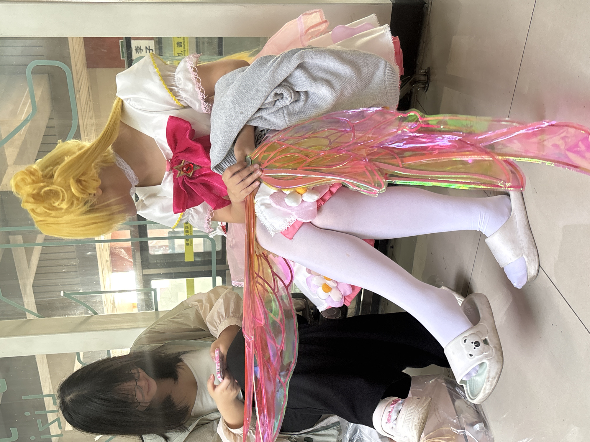 A person cosplaying as Madoka Kaname from the anime series Puella Magi Madoka Magica is sitting down next to another person in Yuzhong District, China. The Madoka cosplayer has blonde hair, a white and pink dress, and white tights, and they are holding pink wings. The other person has black hair and glasses, and they are wearing a beige jacket, a white shirt, and black pants, and they are holding a phone. The location appears to be an indoor space.