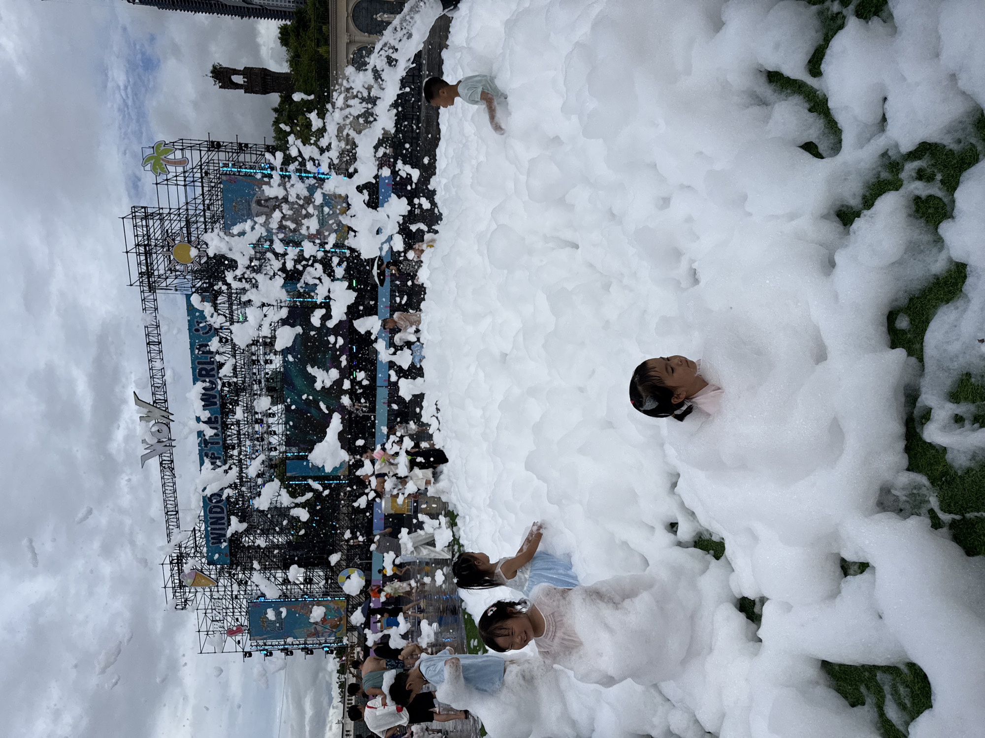In Nanshan District, China, a foam party is in full swing at an outdoor event. A large stage structure, displaying "WINDSOF THE WORLD," dominates the background, adorned with various decorations including an ice cream cone, a sun, and a palm tree. The foreground is filled with a dense layer of white foam, into which several children are playing.
In the foreground, a young girl wearing a light pink dress is partially submerged in the foam, looking to her right with a smile. To her left, another young girl in a light blue dress is seated in the foam, facing away from the camera. Further to the right, a young boy in a light blue t-shirt is also seated in the foam, looking to his right. In the middle ground, another young girl in a light pink top is peeking out from the foam.
Other people, including adults and children, are visible in the background, some standing and others walking. The ground appears to be covered in artificial turf, partially visible beneath the foam. The sky is overcast with clouds. The scene captures a moment of playful enjoyment amidst a lively event.