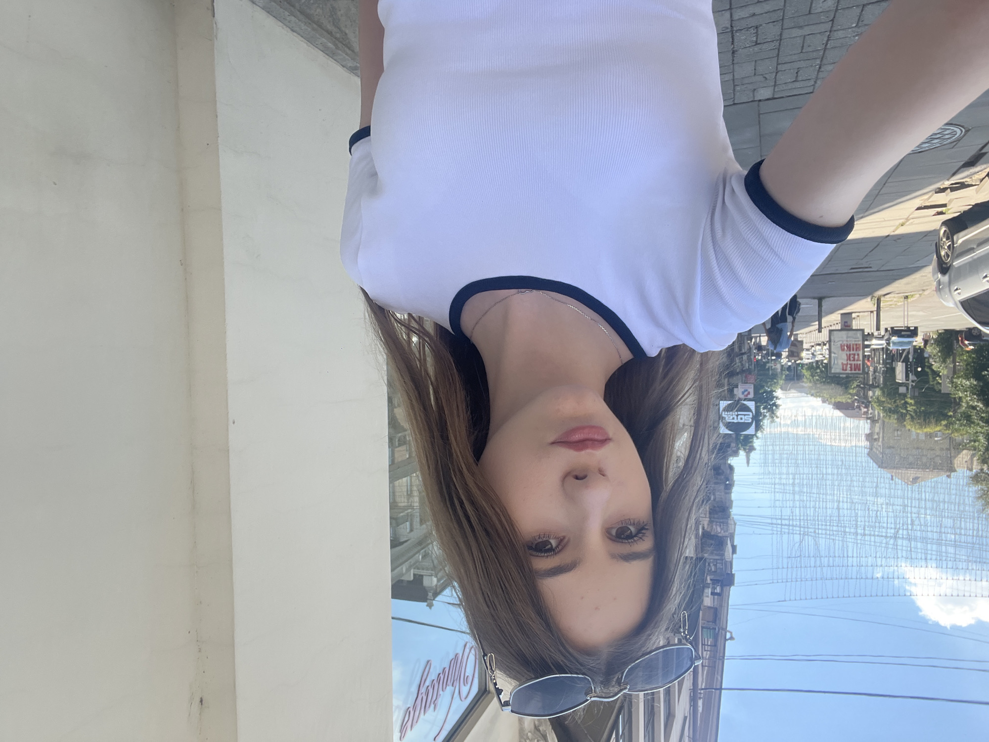 A young woman with long, light brown hair and sunglasses perched on her head takes a selfie on a city street in Dnipro, Ukraine. She is wearing a white t-shirt with dark trim around the collar and sleeves, and a silver necklace with a heart pendant. Behind her, a white building with a window displaying the word "Vintage" is visible. The street is paved with cobblestones, and a sidewalk runs alongside it. In the background, there are trees, cars, buildings, and a clear blue sky with some clouds. The overall scene depicts a casual moment captured outdoors on a sunny day.