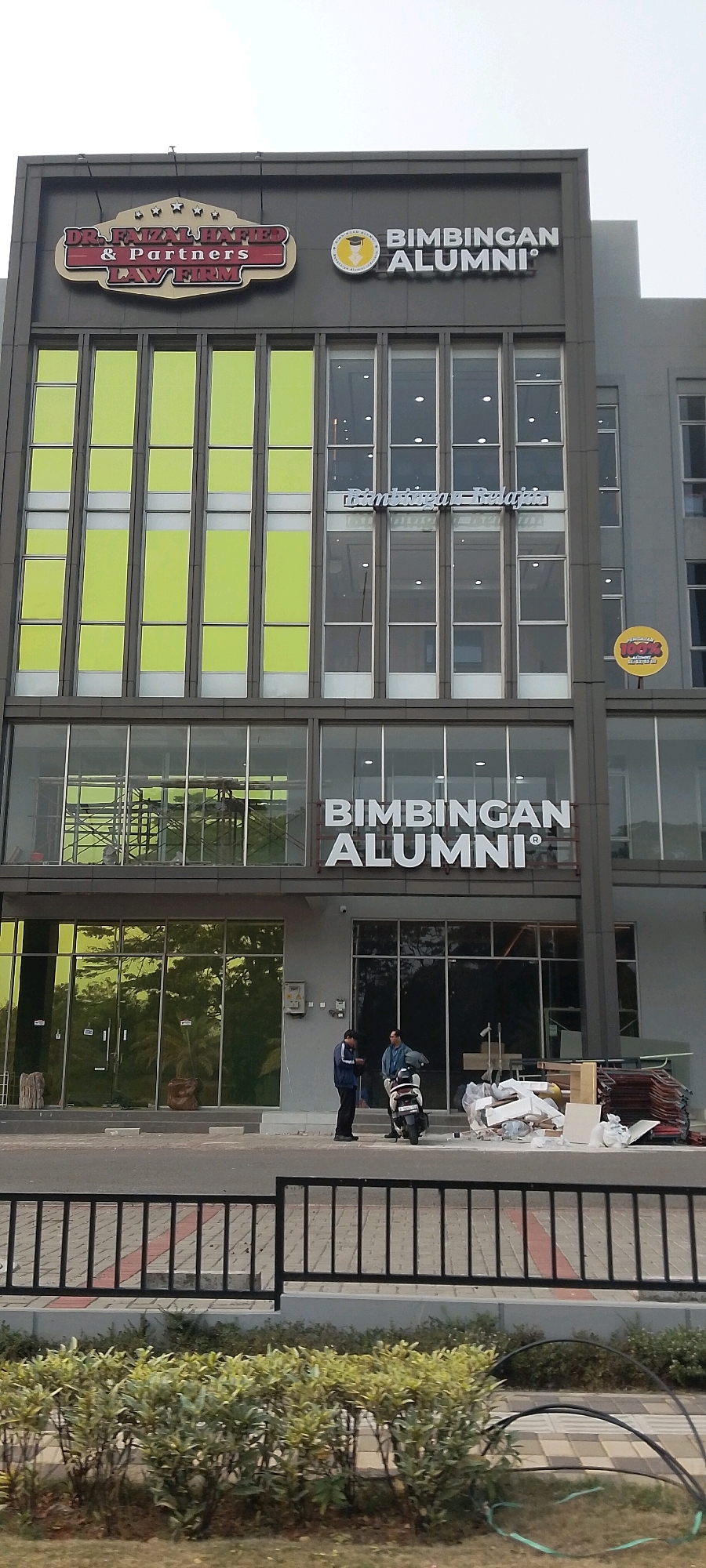 The image shows the exterior of a building in Gunung Putri, Indonesia. The building has a modern architectural style with a dark gray facade and large glass windows. There are two prominent signs on the upper part of the building. The sign on the left reads "DR. FAIZAL HAKIFED & PARTNERS LAW FIRM" in a stylized font with a red and gold color scheme. To the right of this, a circular logo with a graduation cap and scroll is visible, next to the words "BIMBINGAN ALUMNI" in large white letters. Below this, there is another sign that reads "BIMBINGAN ALUMNI" in white, 3D letters.
The lower floors of the building feature large glass windows, some with a greenish tint and others reflecting the surrounding environment. In front of the building, a black metal fence runs horizontally. Beyond the fence, there are low green bushes and a paved walkway. On the ground level of the building, two men are standing near a scooter. One man, wearing a blue jacket and dark pants, is facing away from the camera and looking down at something in his hand. The other man, in a dark jacket and lighter pants, is standing next to him, also looking down. A scooter is parked between them. To the right of the men, there is a pile of construction debris, including what appears to be packaging materials and some roofing tiles. The sky in the background is overcast.